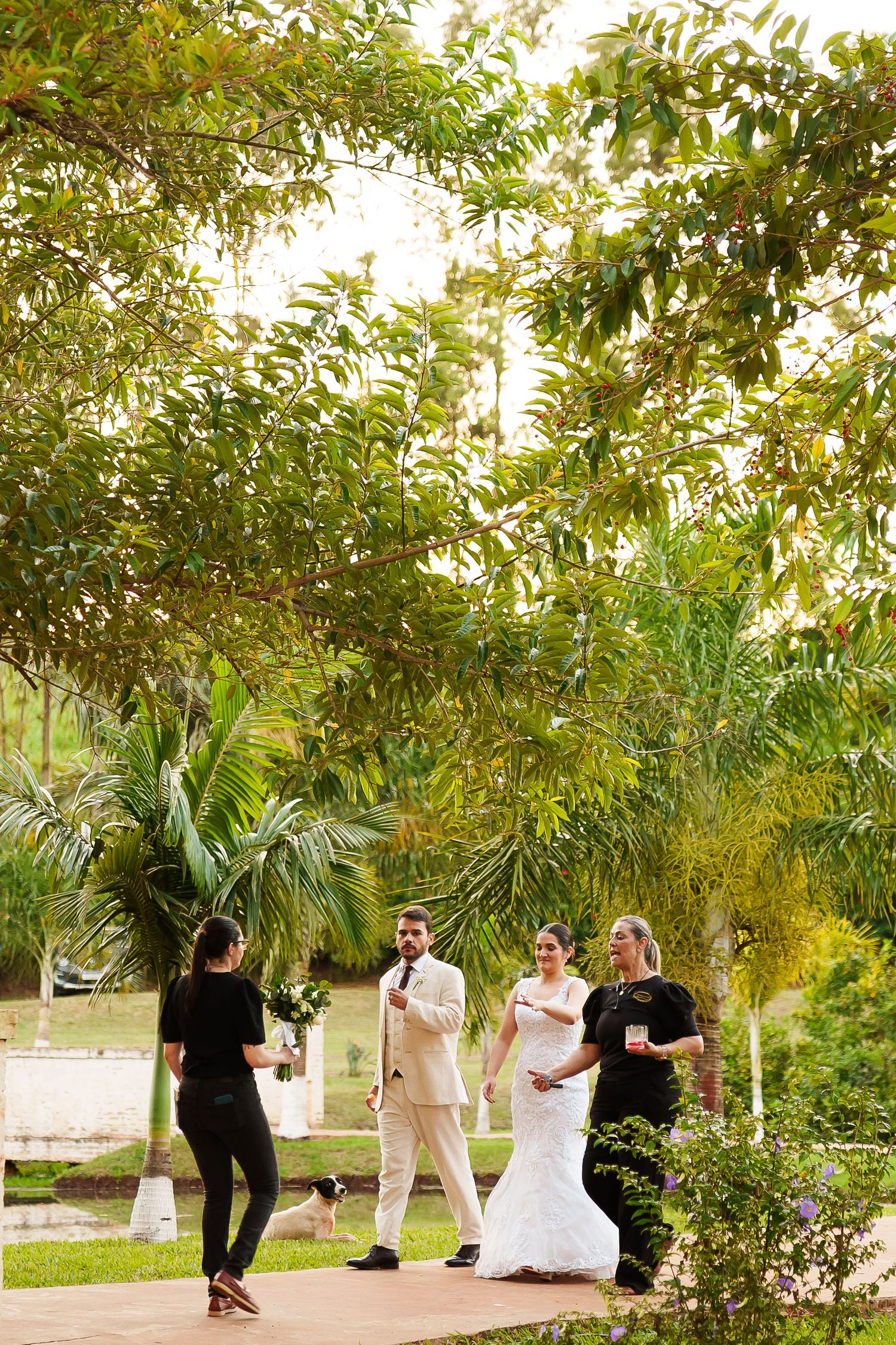Bride in white gown and groom in beige suit stand with guests in a lush garden setting during an outdoor wedding.