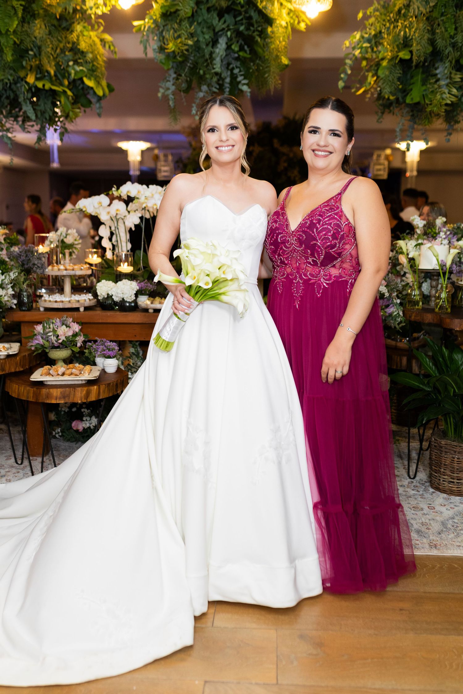 Bride in white ball gown holding bouquet poses with woman in magenta dress at elegant floral wedding reception.
