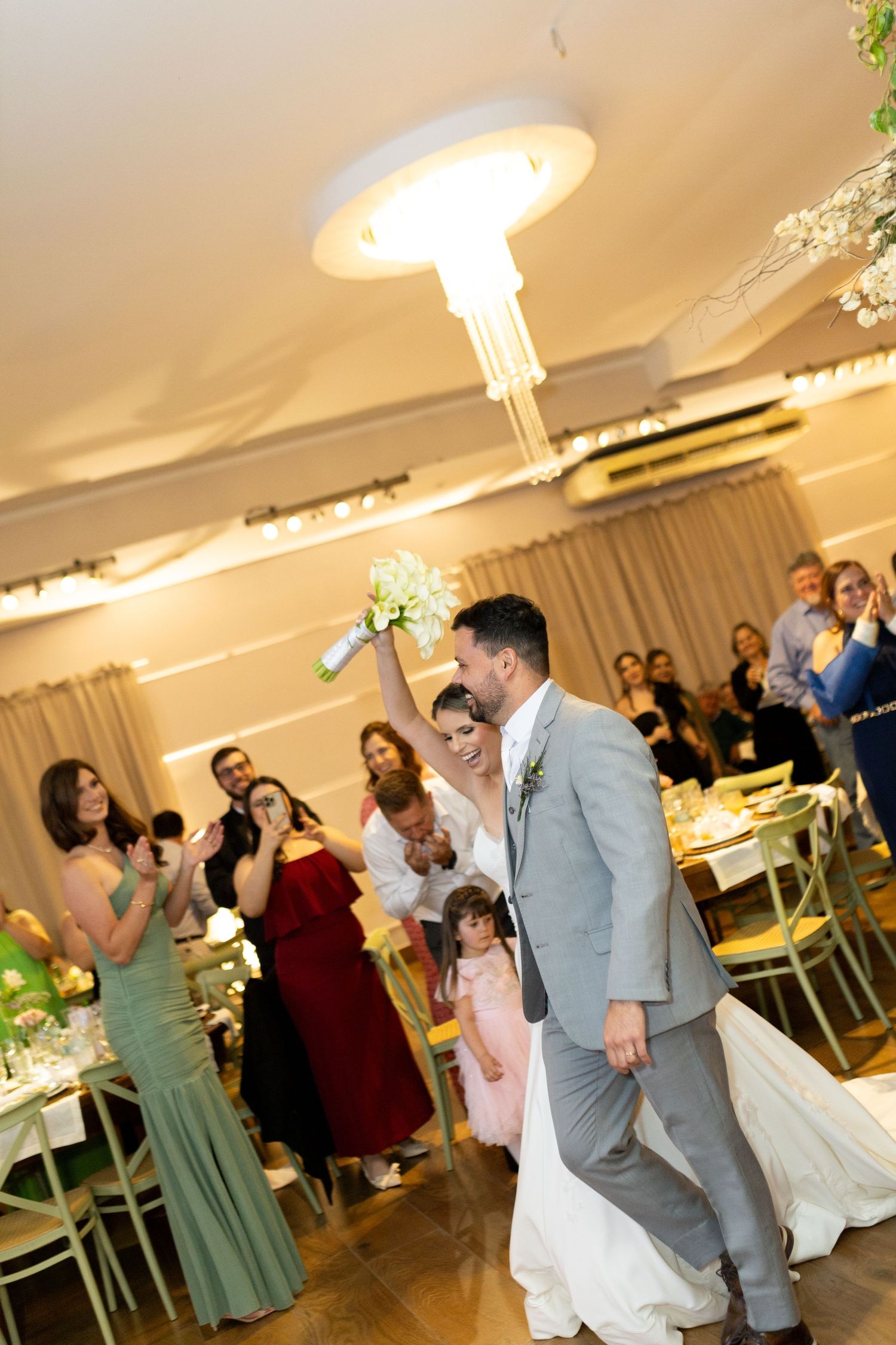 Bride and groom enter wedding reception as guests applaud, bride raising bouquet in elegant venue.