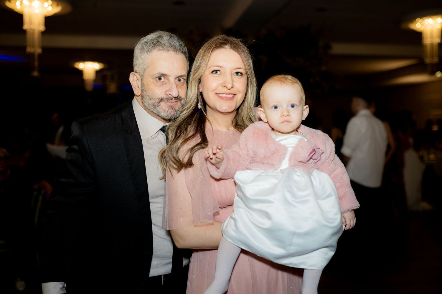 A couple in formal attire smiles while holding a baby in a white christening gown at an elegant event.