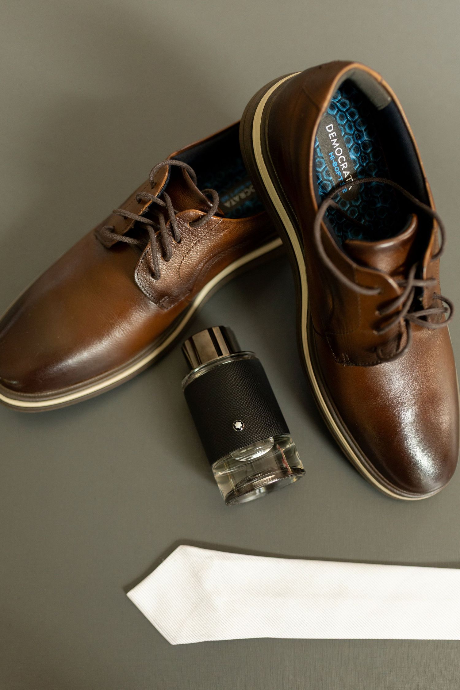 Brown leather dress shoes with white tie and Montblanc cologne arranged on gray surface.