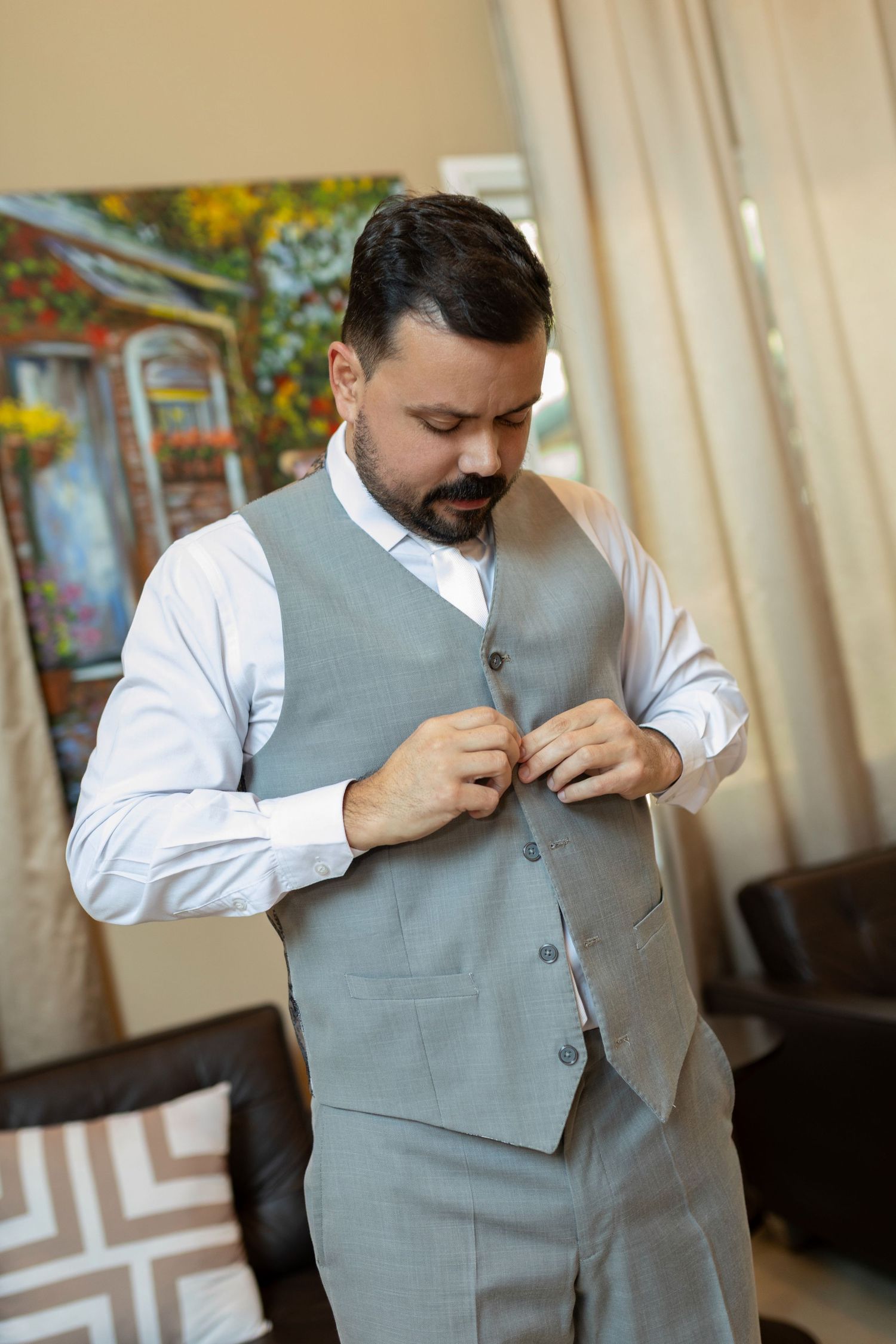 A man in a gray vest and white dress shirt buttoning his waistcoat while getting ready indoors.