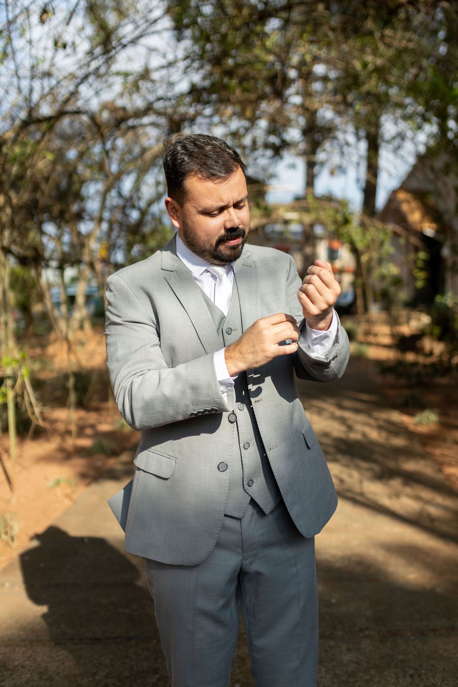 Man in gray three-piece suit adjusting cufflinks outdoors among trees on a sunny day.