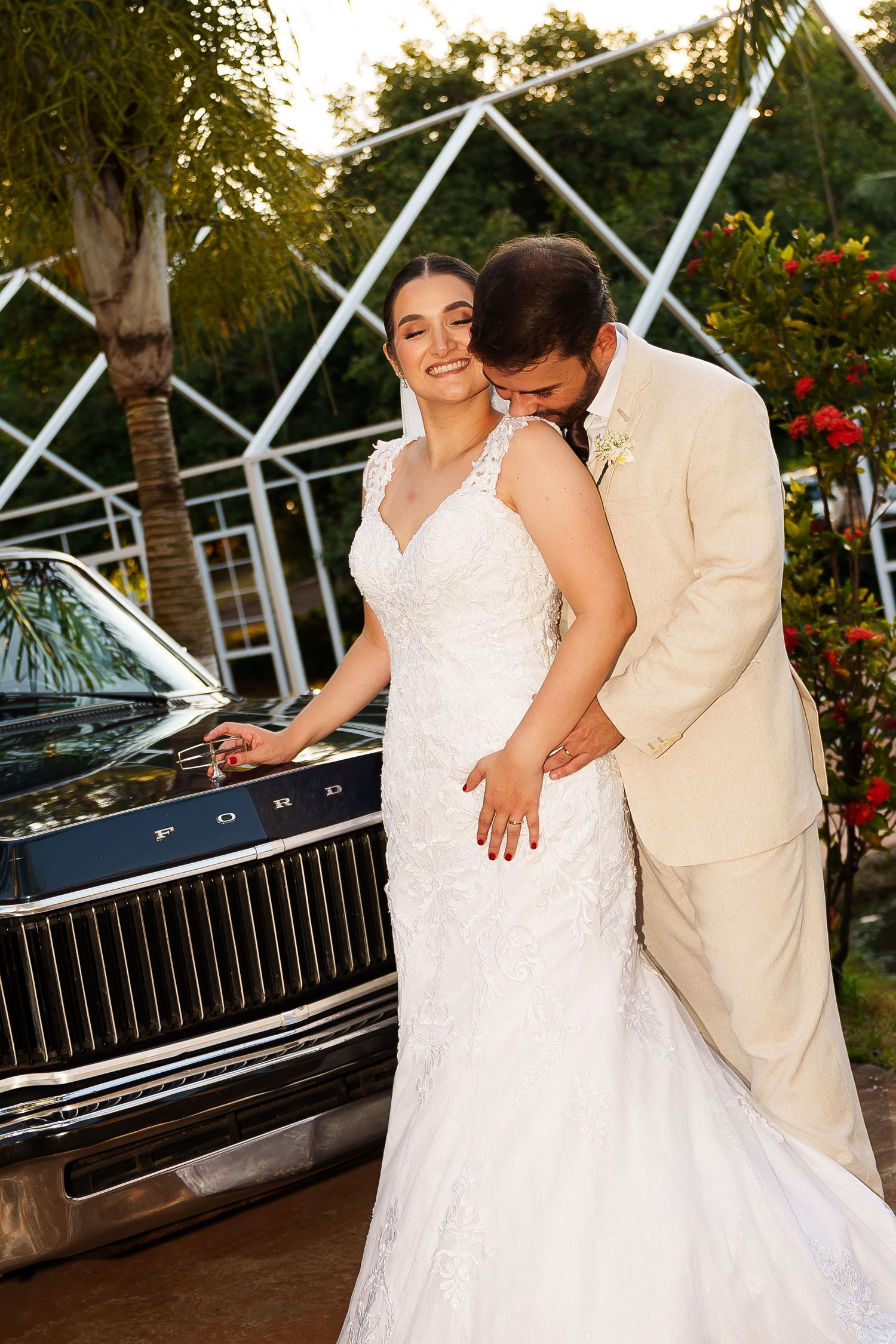 Bride in lace gown and groom in beige suit pose beside a classic Ford car at an outdoor wedding venue.