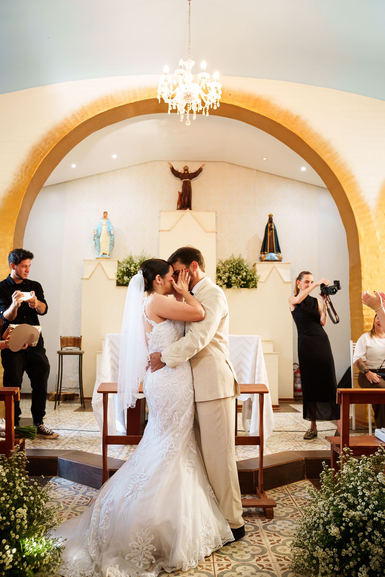 Bride and groom share first kiss at church altar, surrounded by floral arrangements and religious statues.