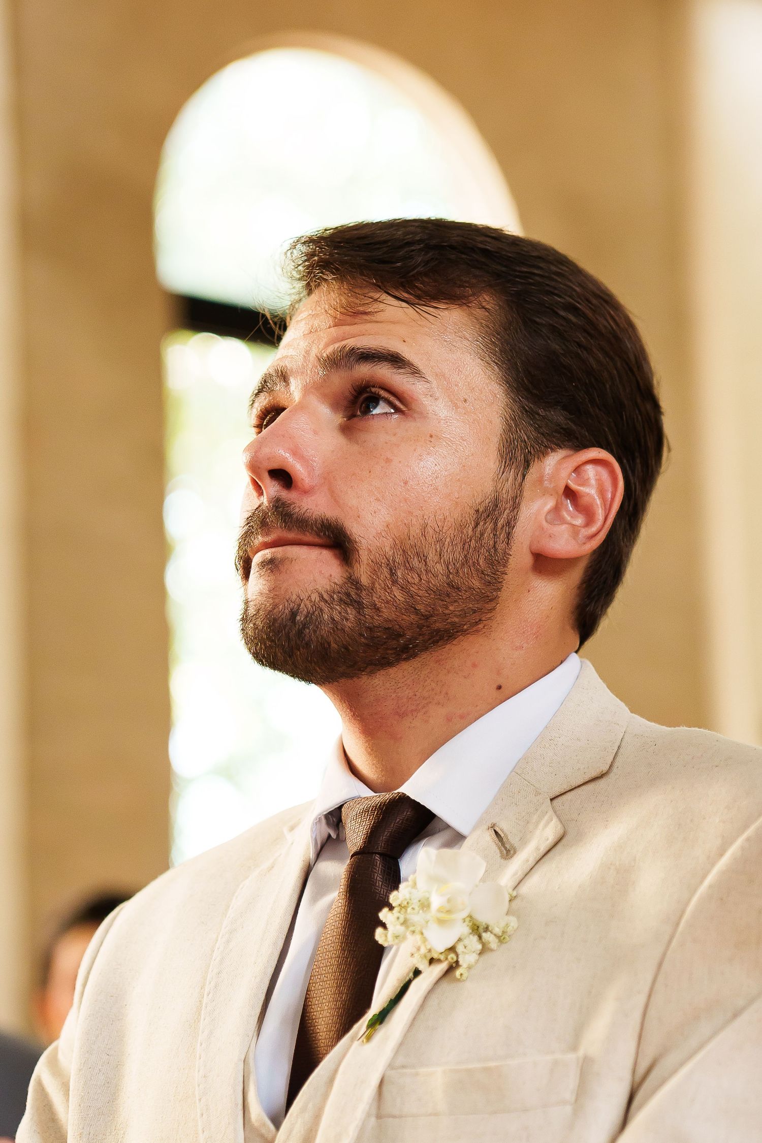 A bearded groom in a cream suit with white boutonniere gazes upward emotionally during a wedding ceremony.