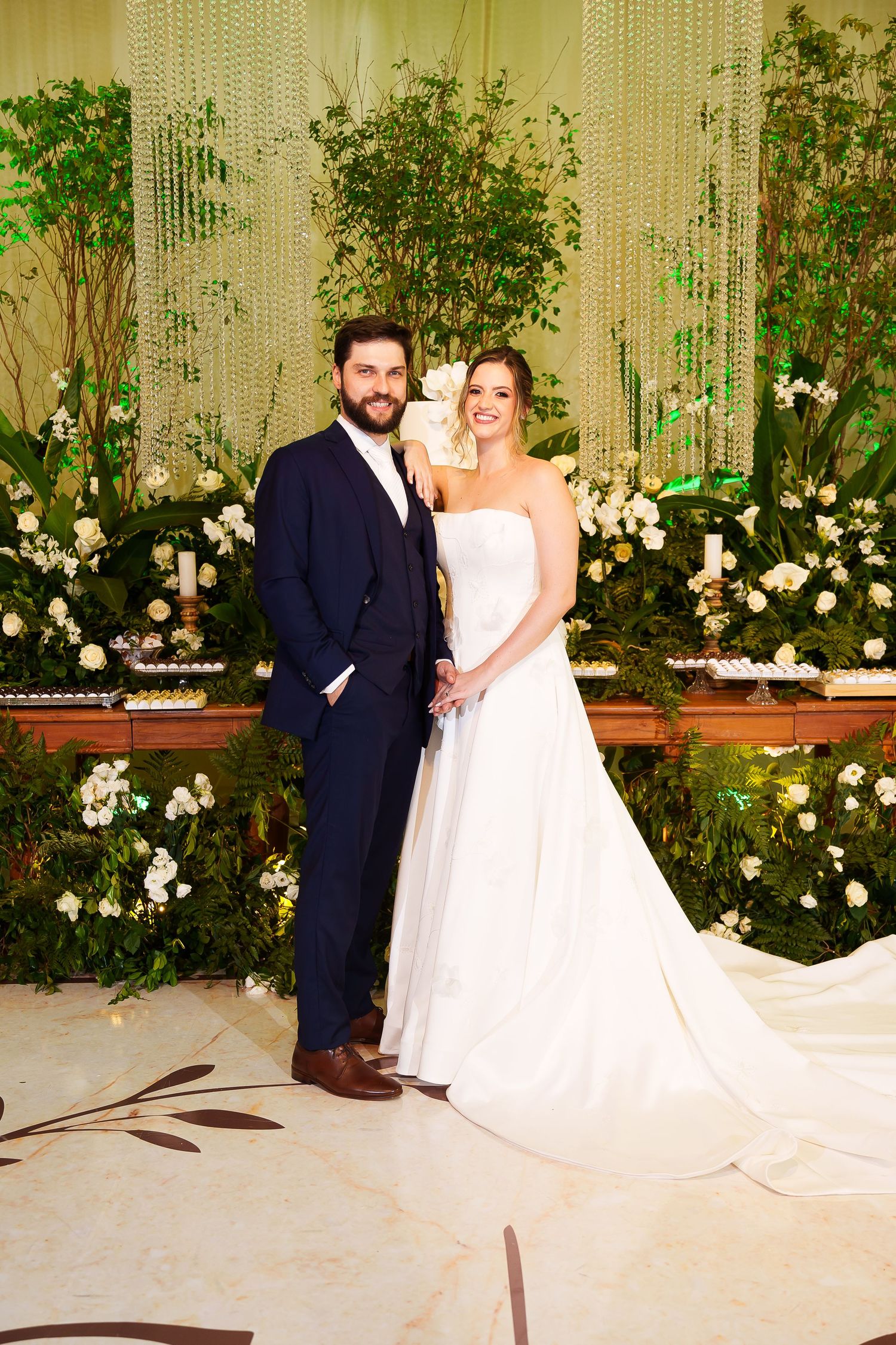 Bride and groom posing together at wedding reception with lush greenery and white floral arrangements backdrop.