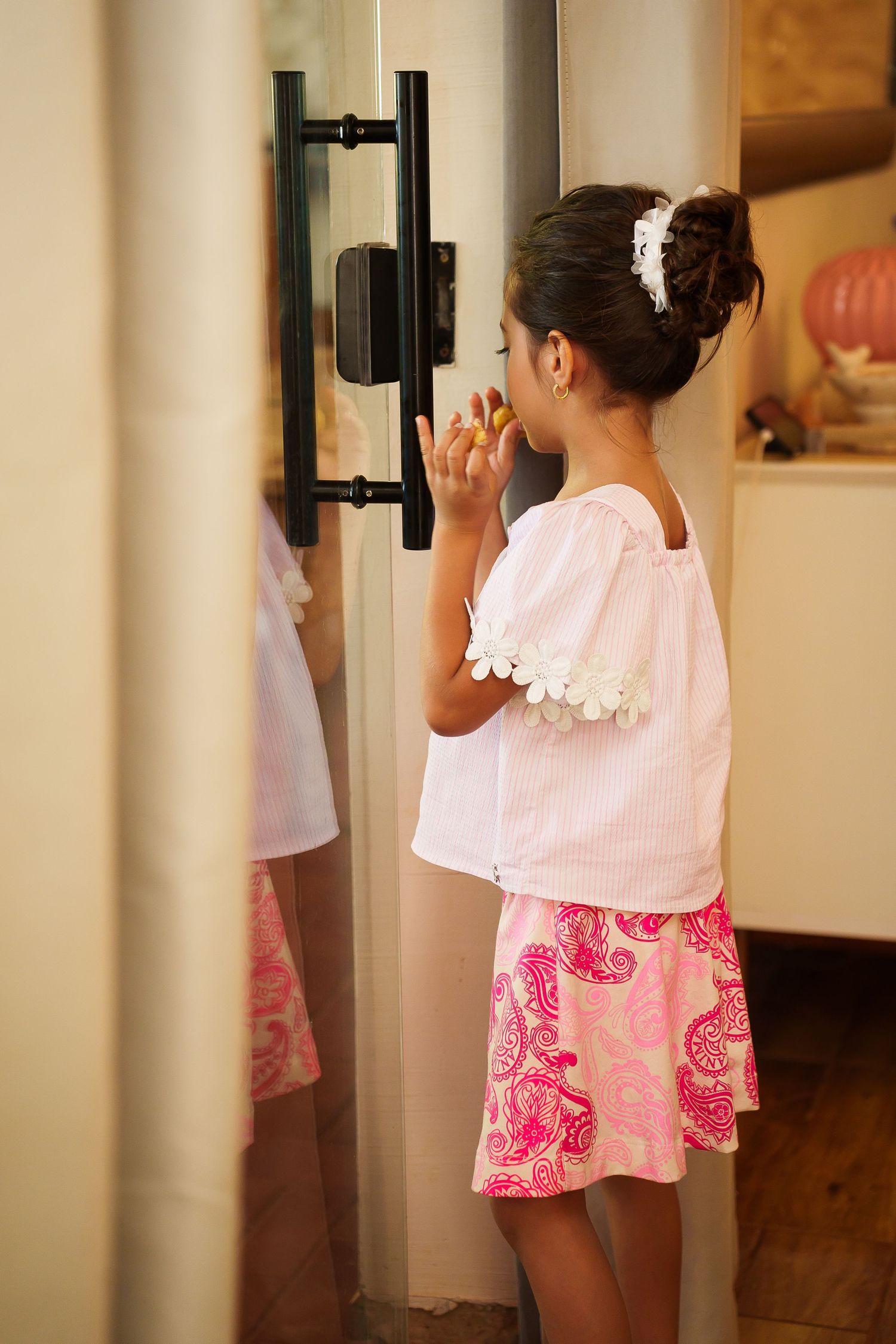 Young girl in white top and pink skirt eating a snack while standing in front of a refrigerator.