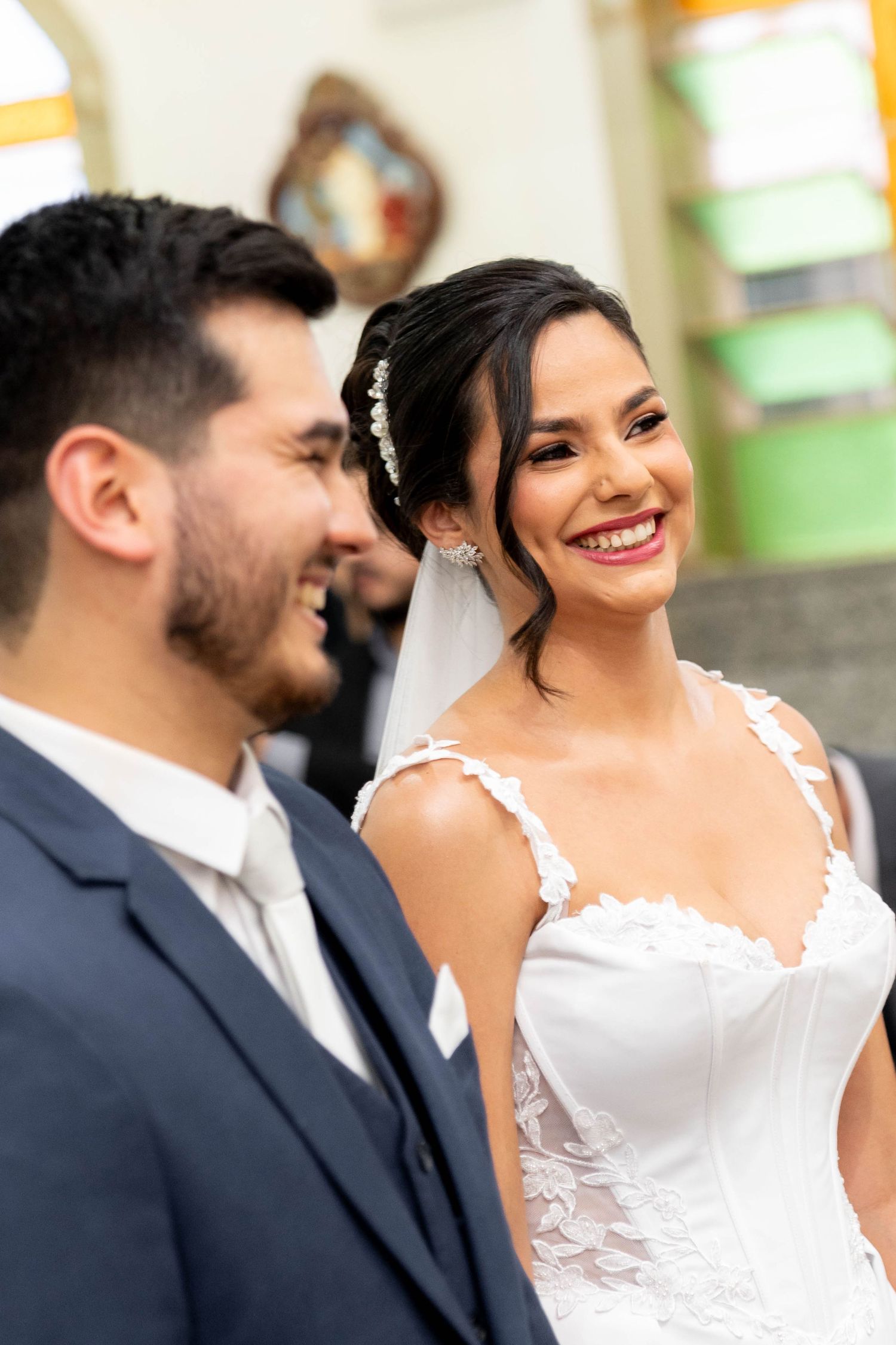 A smiling bride in a white lace gown and veil stands beside a groom in a navy suit during a church wedding ceremony.
