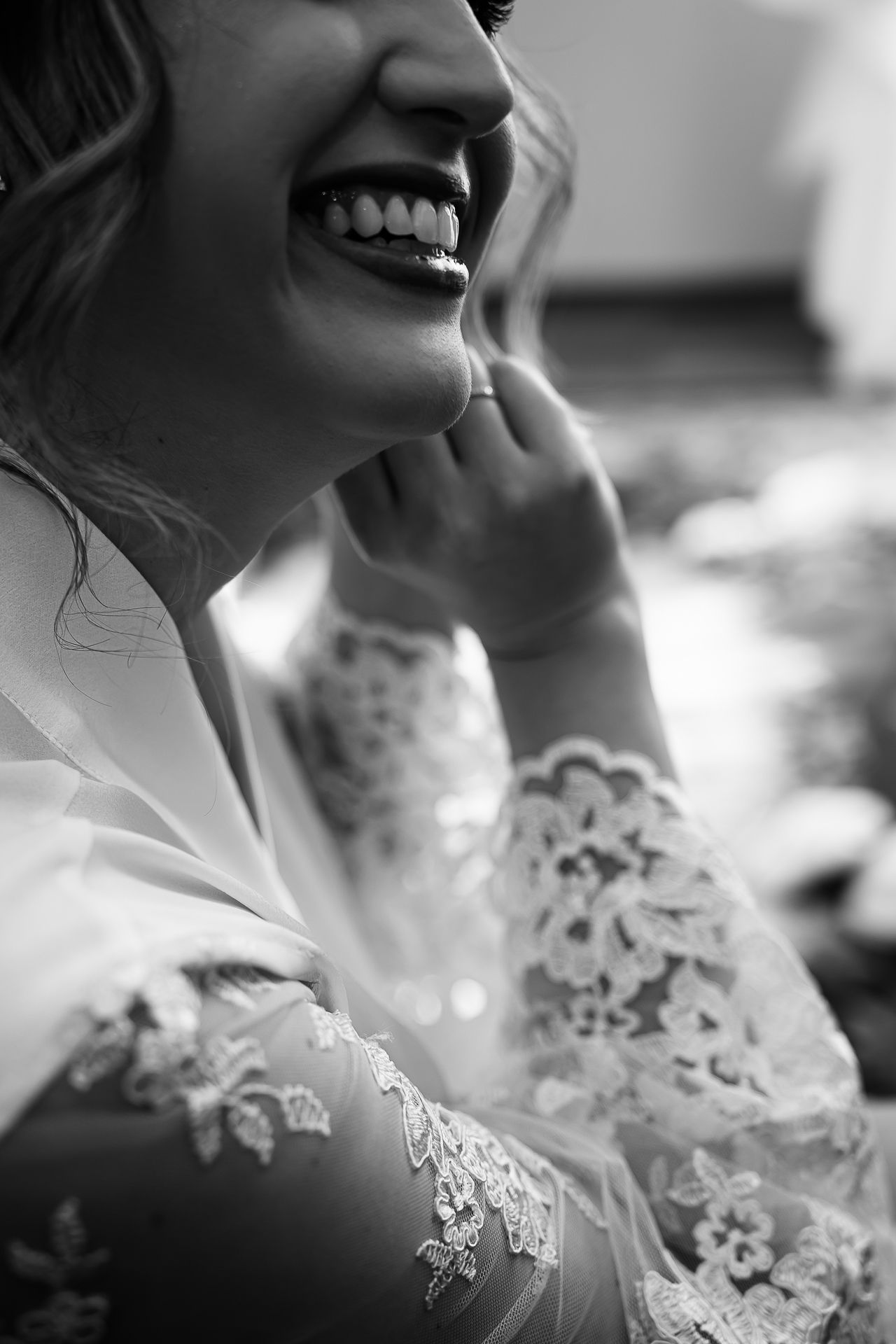 Black and white photo of a laughing bride in lace wedding dress touching her ear.