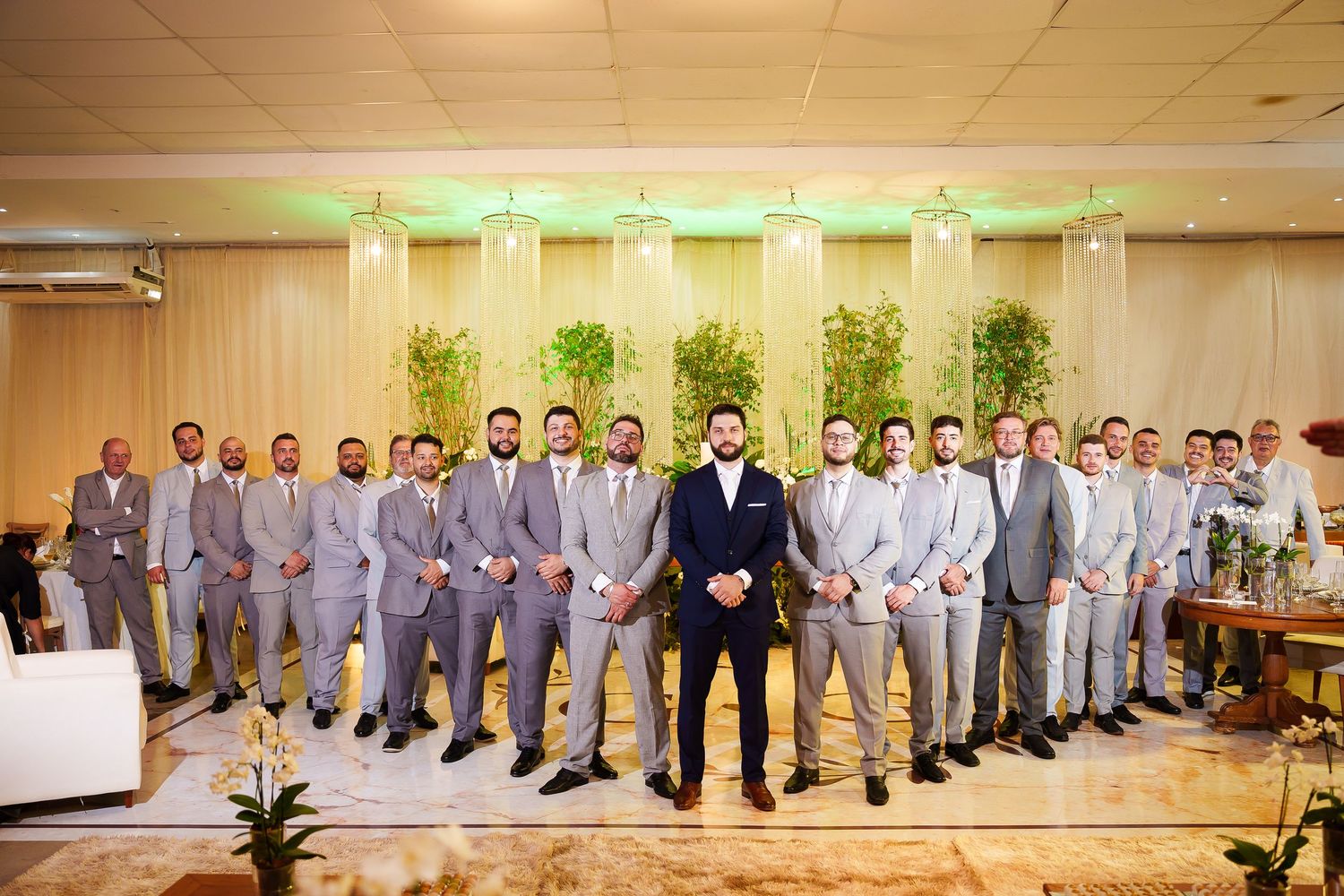 Groom in navy suit stands with large groomsmen group in gray suits at elegant wedding venue with green lighting.
