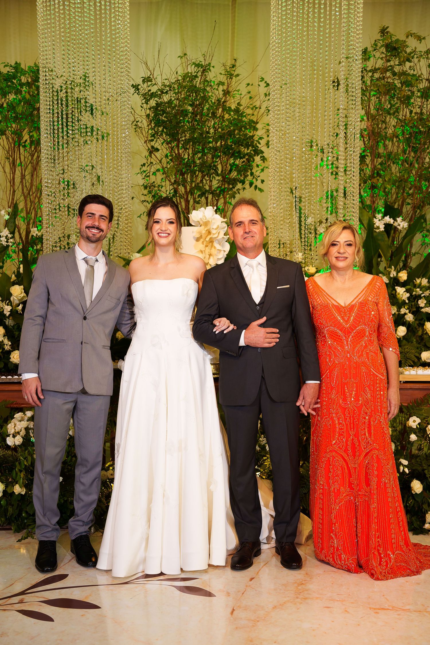 Wedding party of four posing elegantly, bride in white gown, groom in tuxedo, guests in gray suit and orange dress.