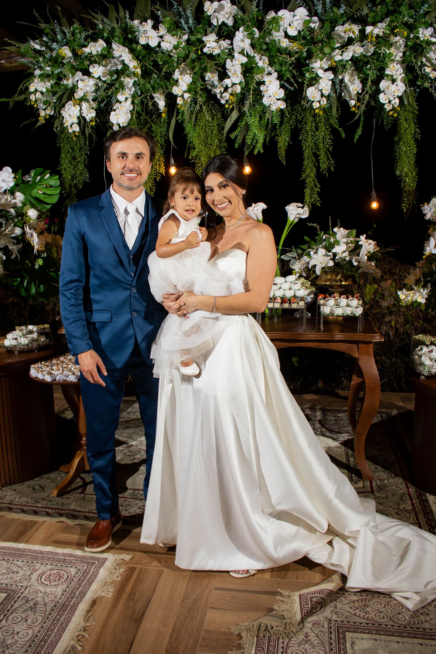 Bride in white gown and groom in blue suit pose with baby girl at elegant floral wedding reception.
