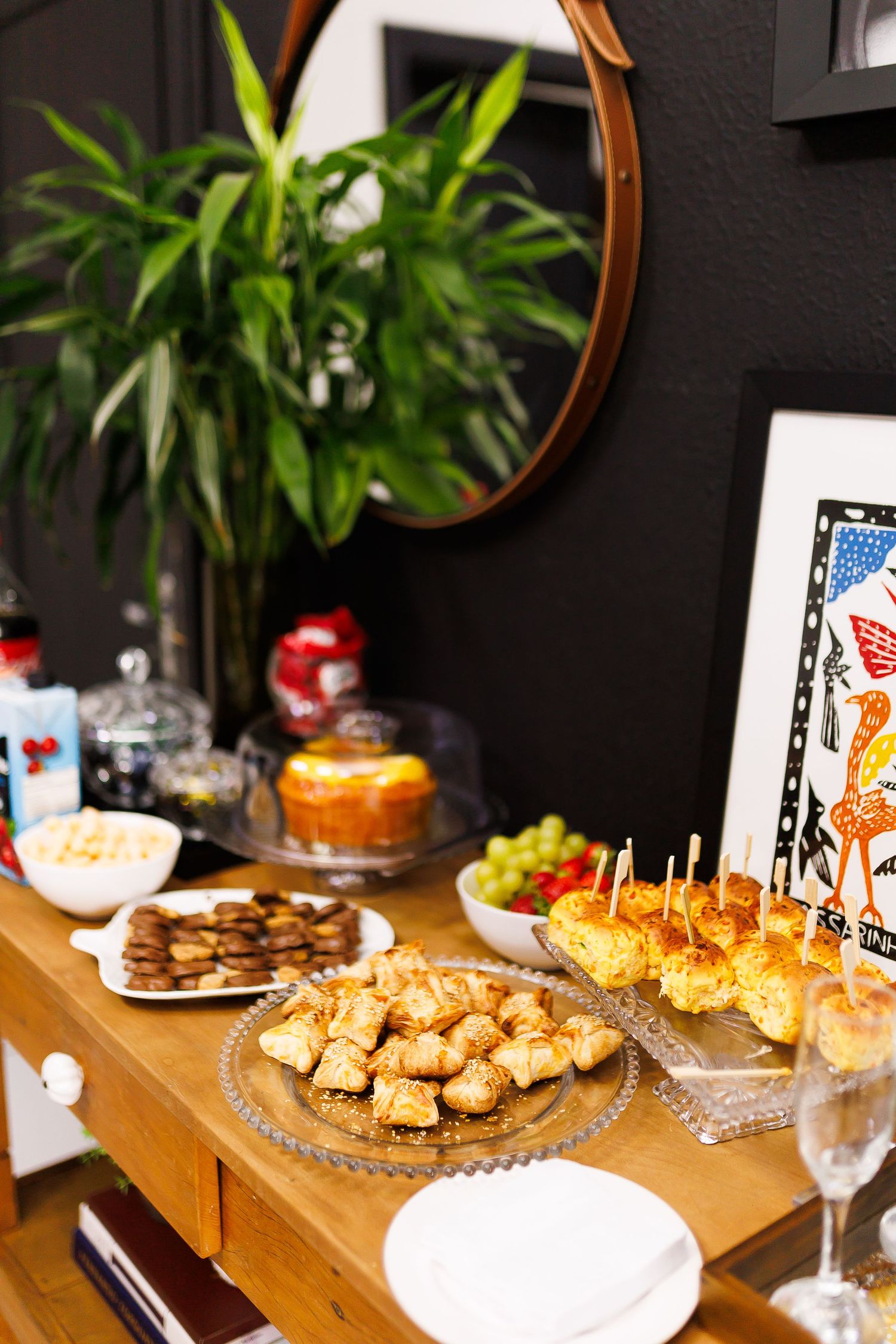 A buffet table with pastries, fruit, and appetizers displayed on glass plates beside a plant and round mirror.