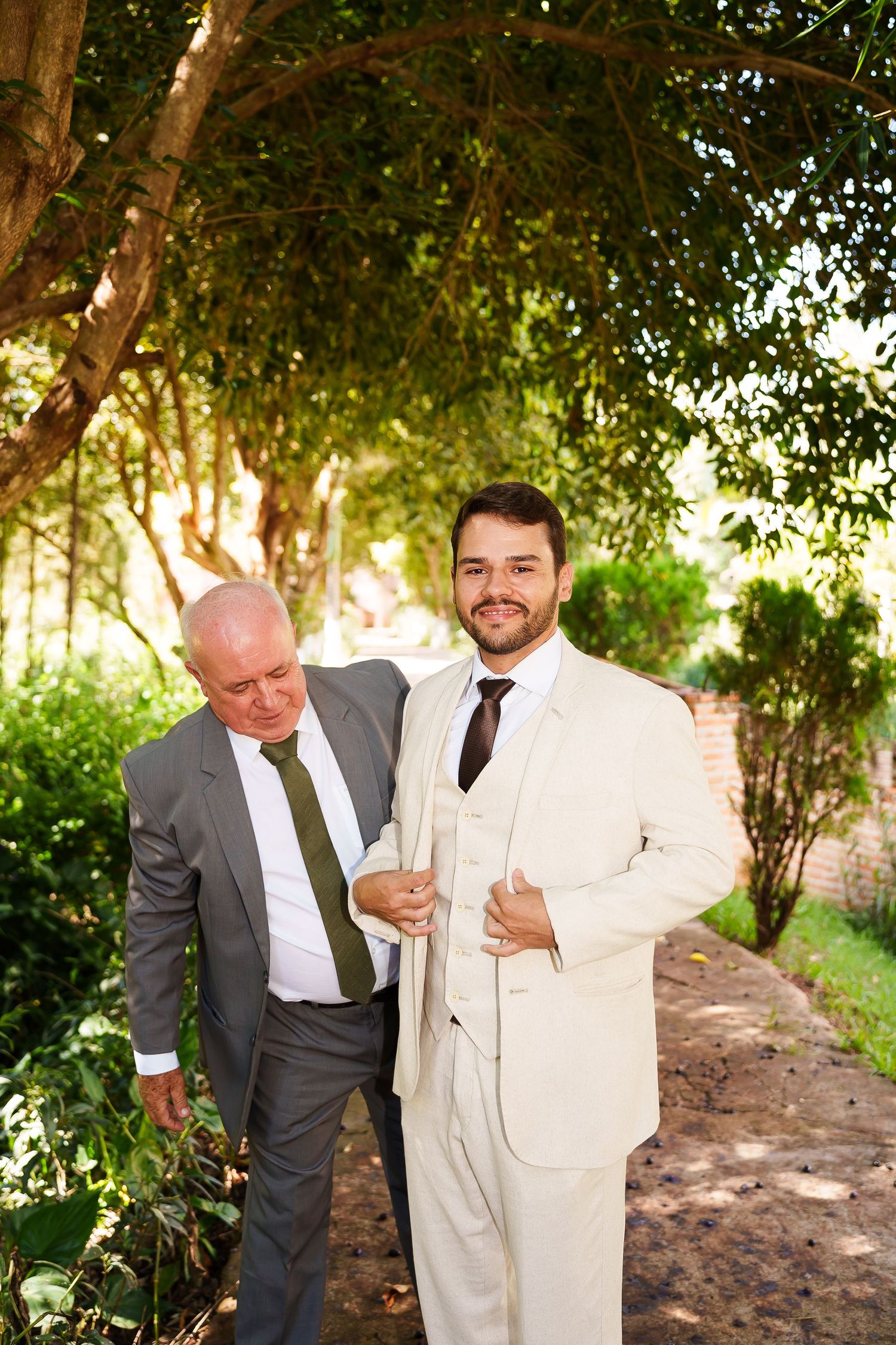 Two men in suits pose outdoors under green trees, smiling at a wedding celebration.