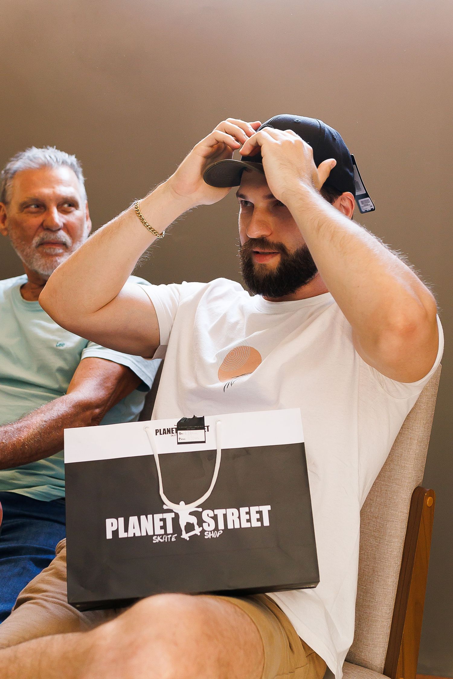 Man trying on a black cap holding a Planet Street shopping bag while seated indoors.