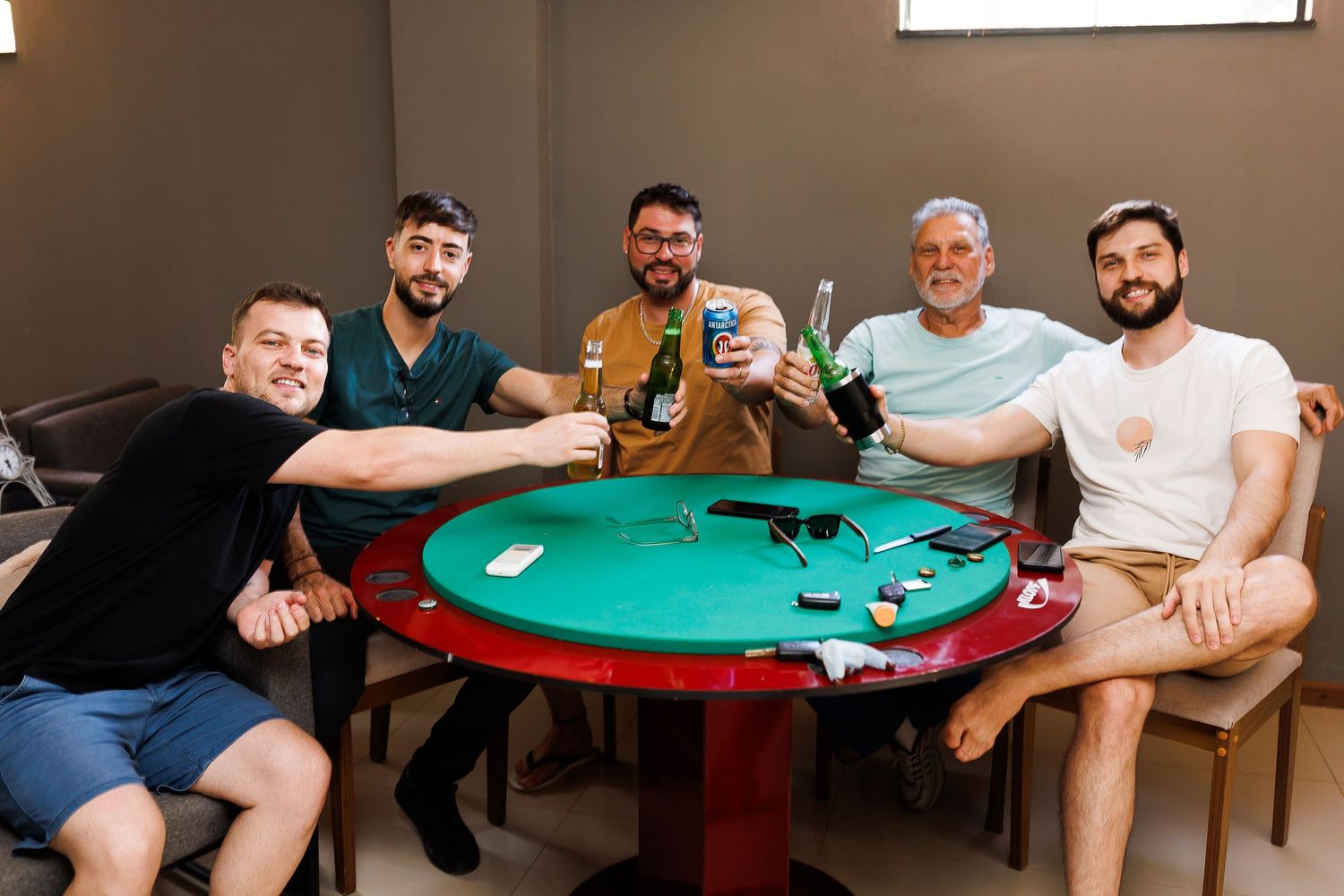 Five men smiling and toasting drinks around a green poker table in a casual indoor setting.