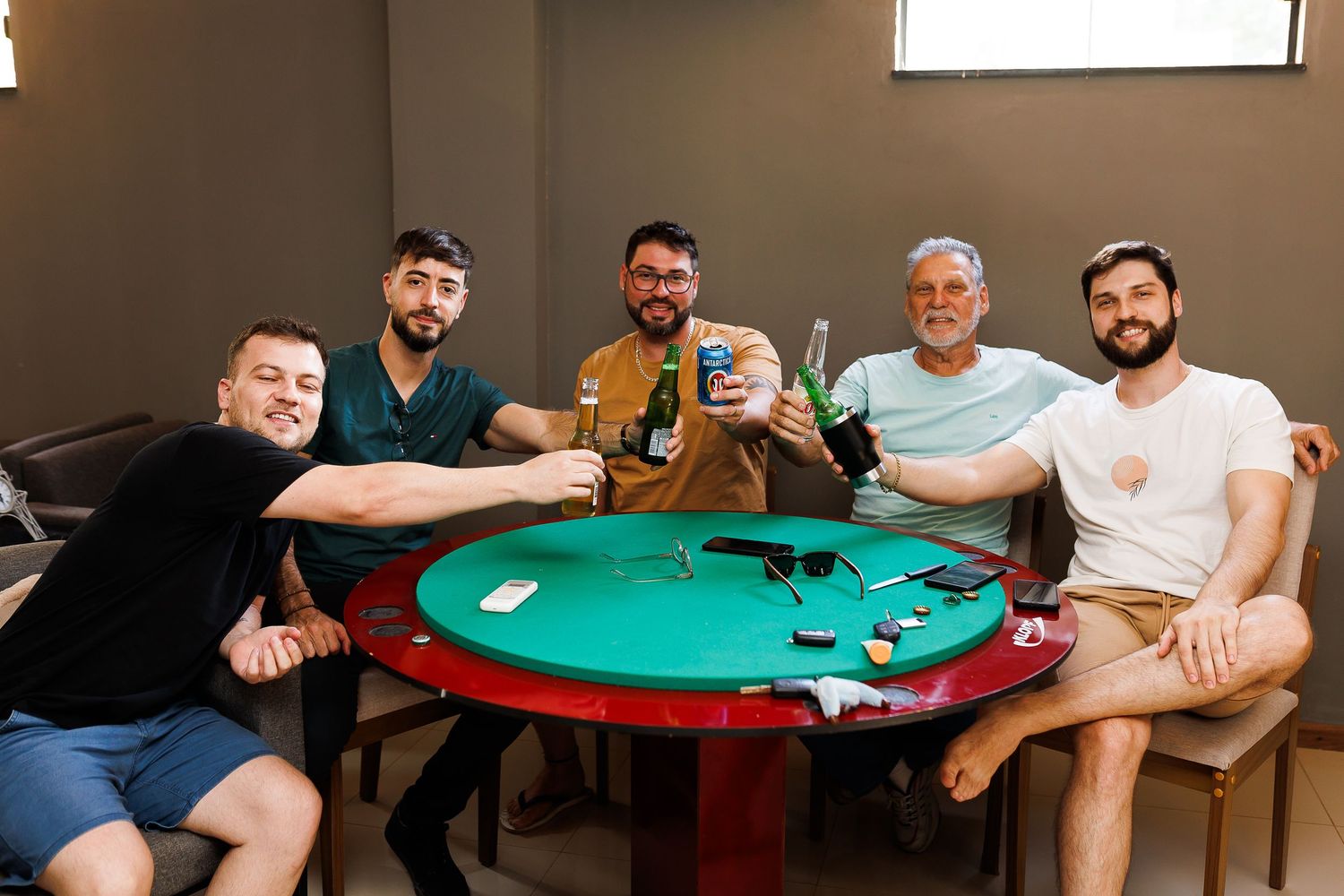 Five men smiling and toasting with beer bottles around a round green poker table indoors.