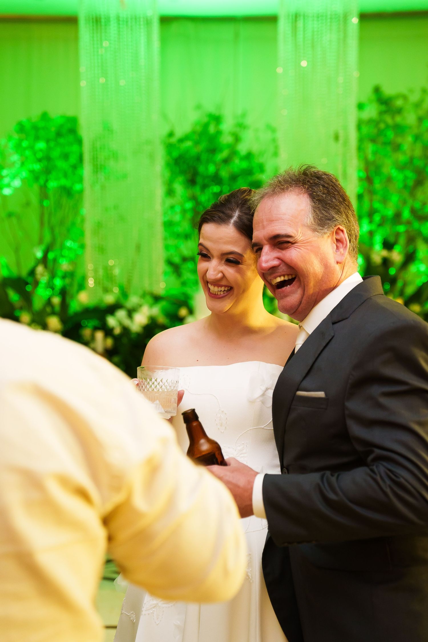 Bride and groom laughing joyfully at their wedding reception with green floral backdrop.