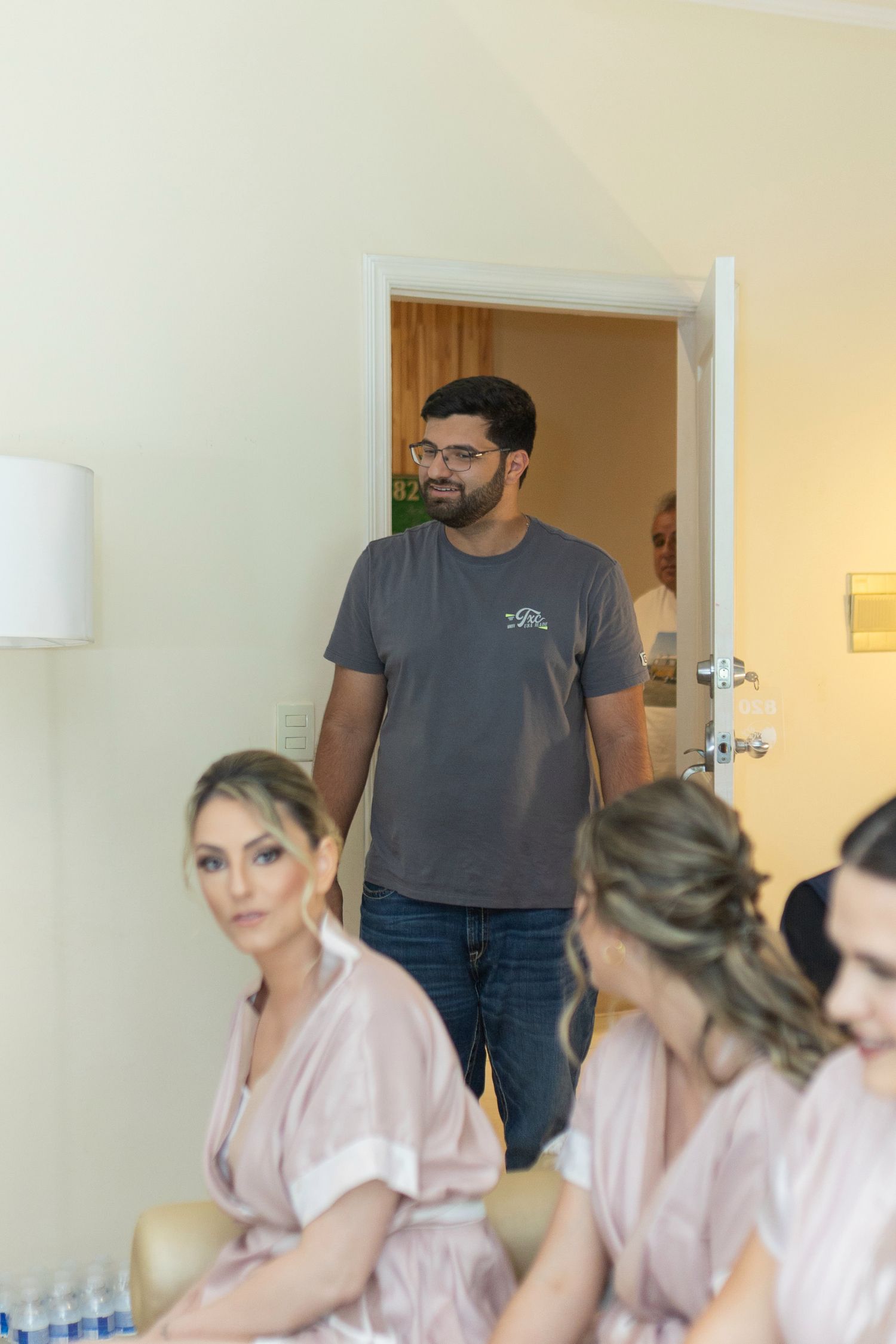 Man in gray t-shirt stands in doorway watching women in pink robes getting ready for a wedding.