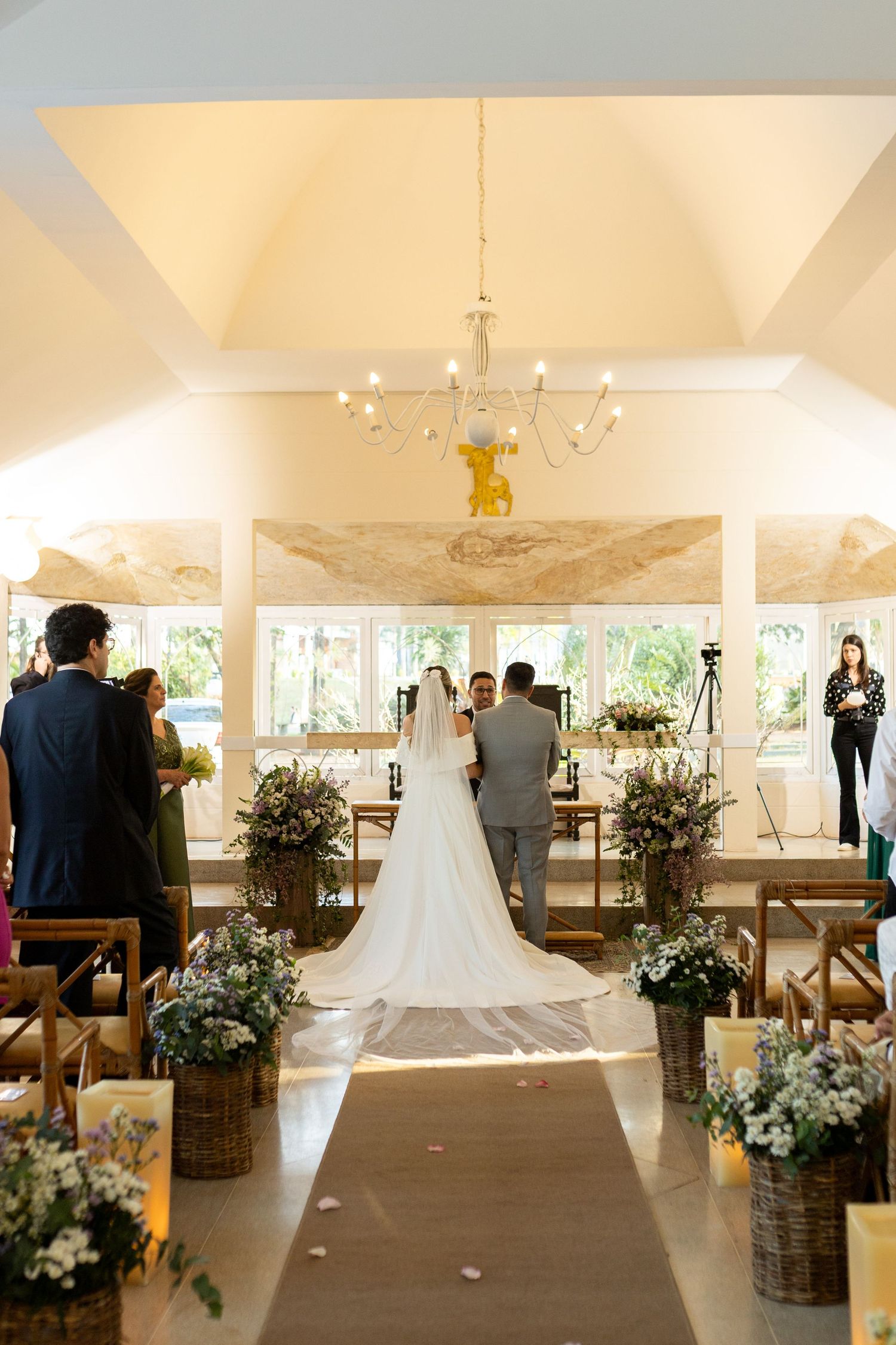 Bride walks down floral-lined aisle in elegant chapel wedding ceremony with chandelier and long veil.