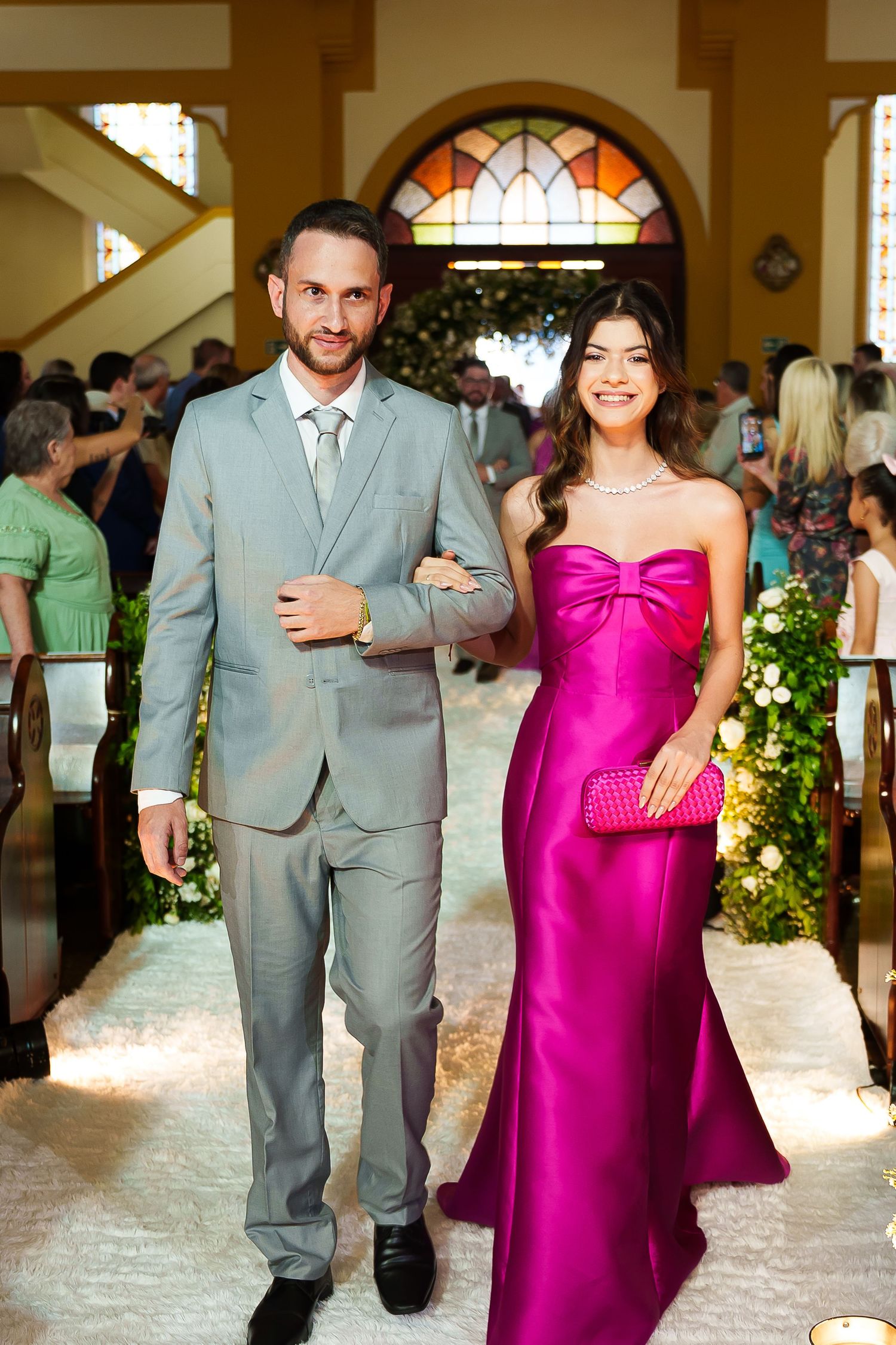 Elegant couple walking down church aisle, man in gray suit, woman in strapless magenta gown with pearl necklace.