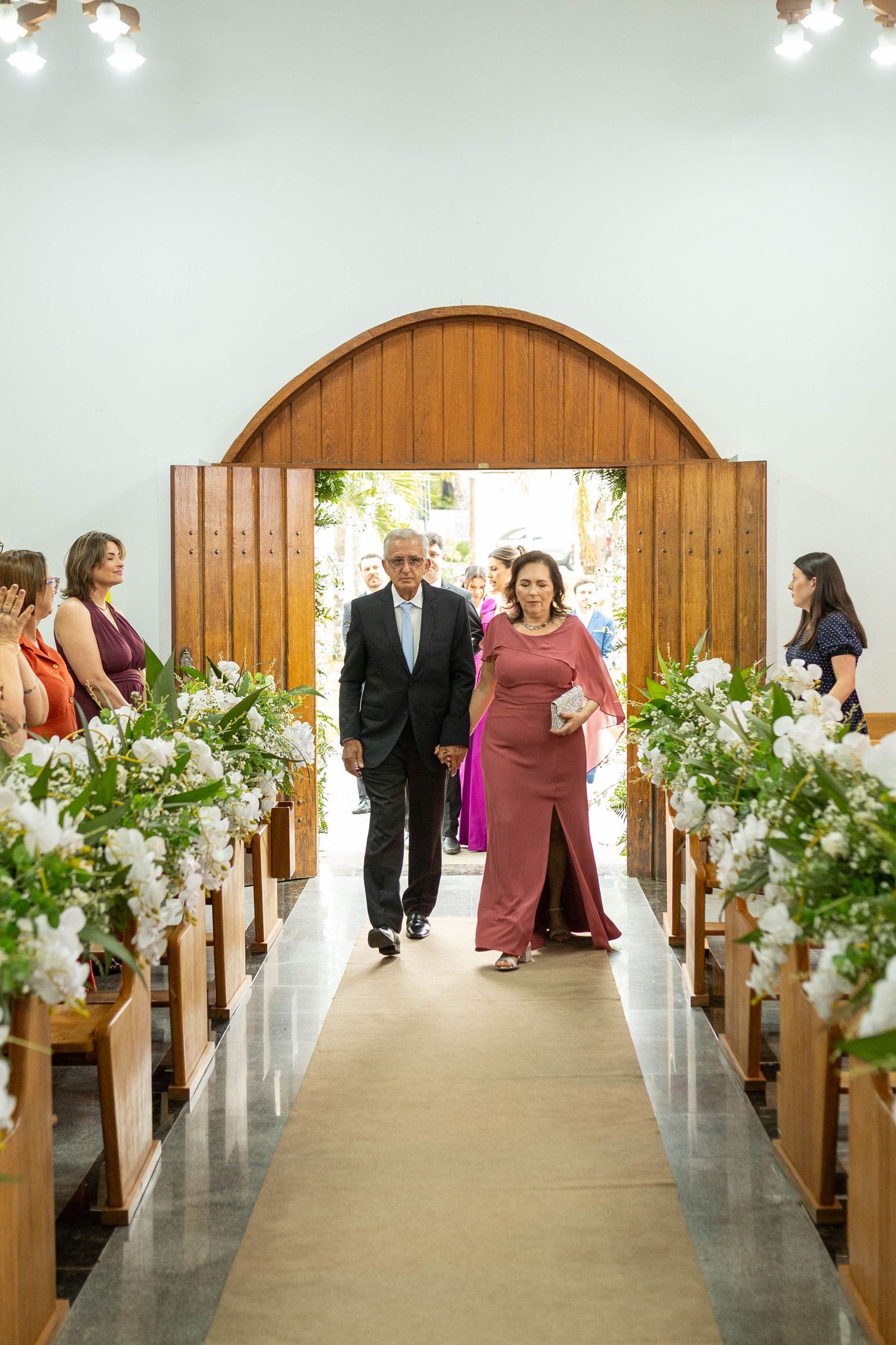 Elegant couple walking down a flower-adorned church aisle during a wedding ceremony, man in suit and woman in pink gown.