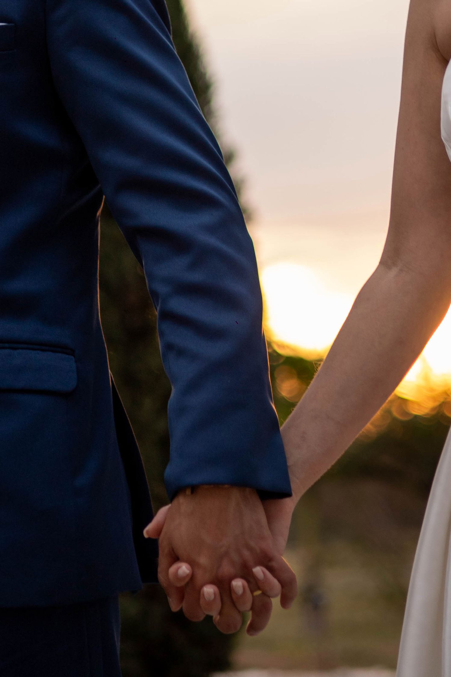 Bride and groom holding hands at sunset, man in blue suit and woman in white dress, romantic wedding moment outdoors.
