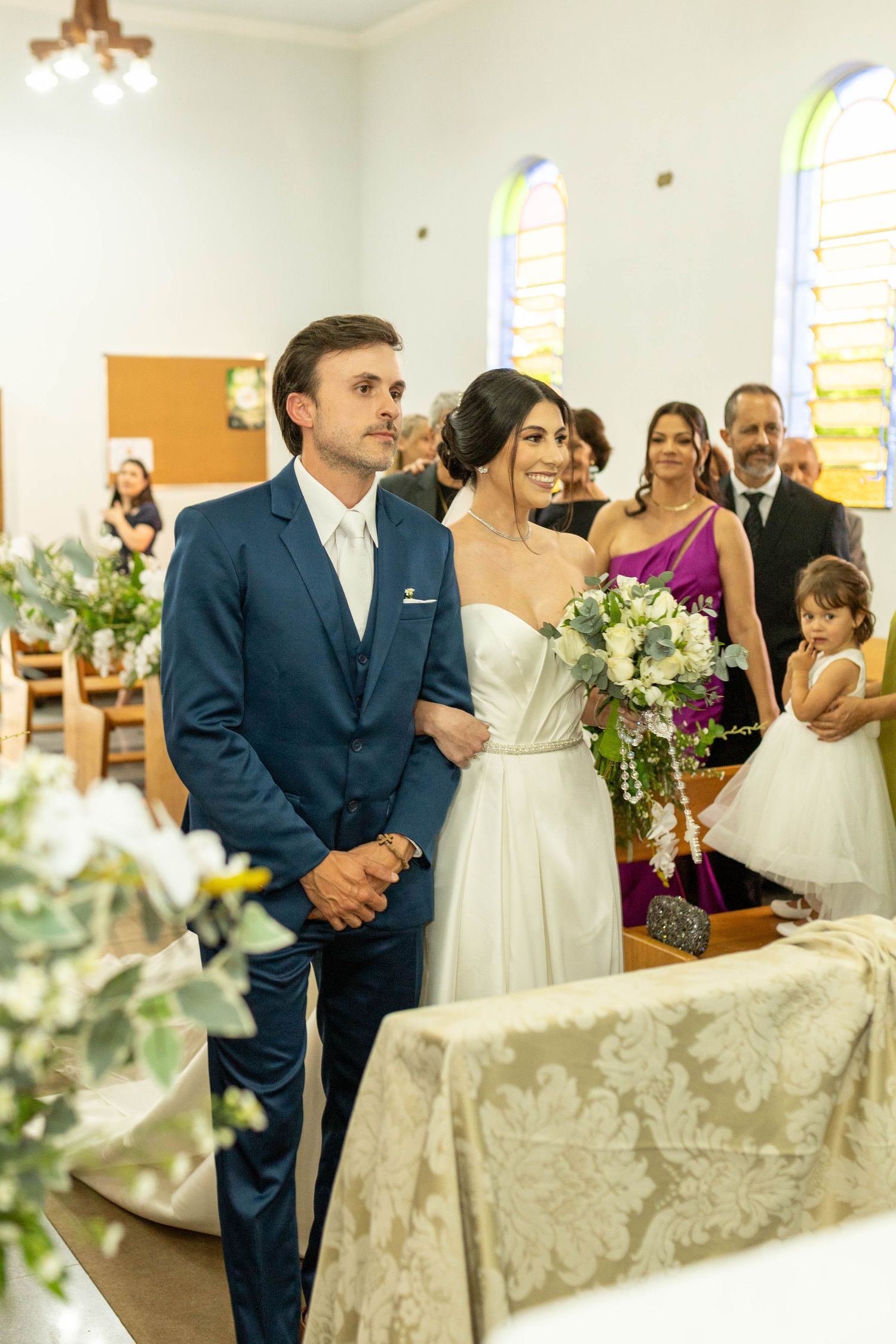 Bride and groom stand at the altar in a church wedding ceremony, she holds a white bouquet, guests seated behind them.