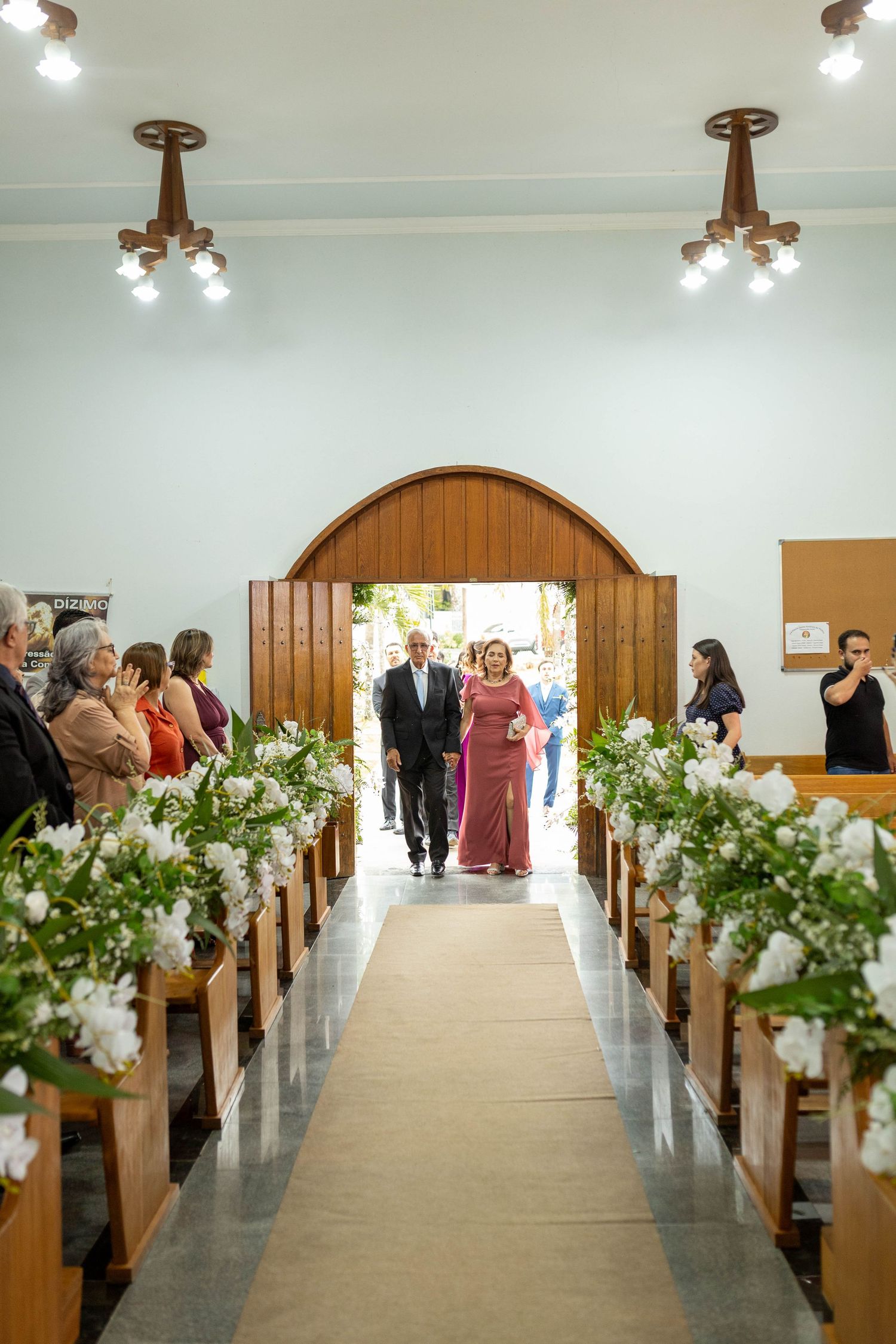 A couple walks down a flower-adorned church aisle during a wedding ceremony, with guests seated on wooden pews.