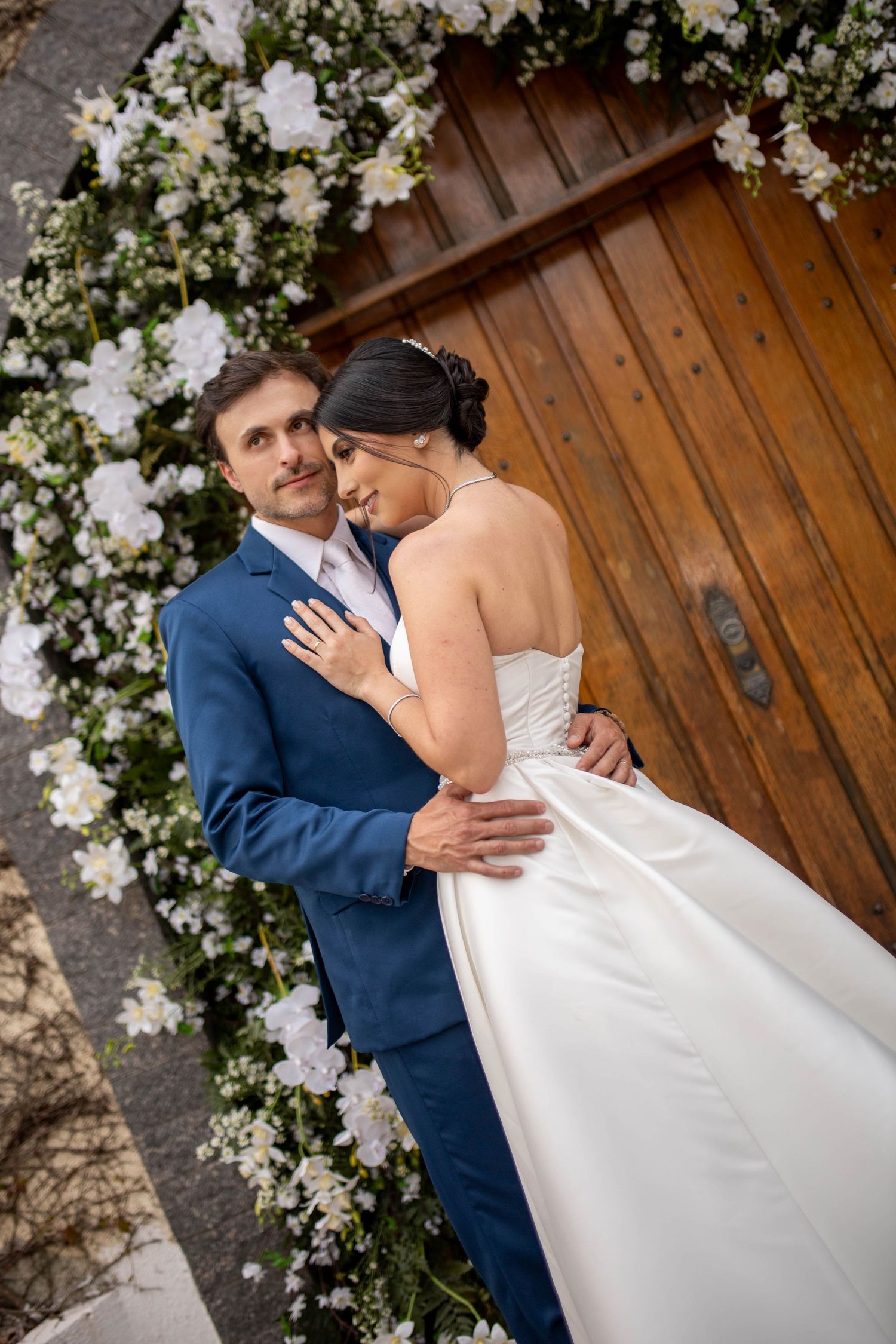 A bride and groom embrace before a floral arch with white flowers at a wooden church door on their wedding day.