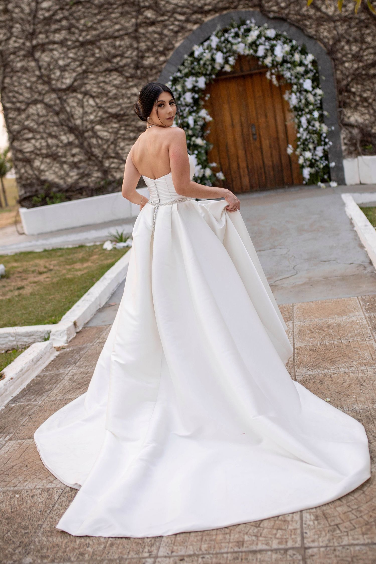 Bride in strapless white satin ballgown with large bow and cathedral train, standing before floral arch and wooden door.