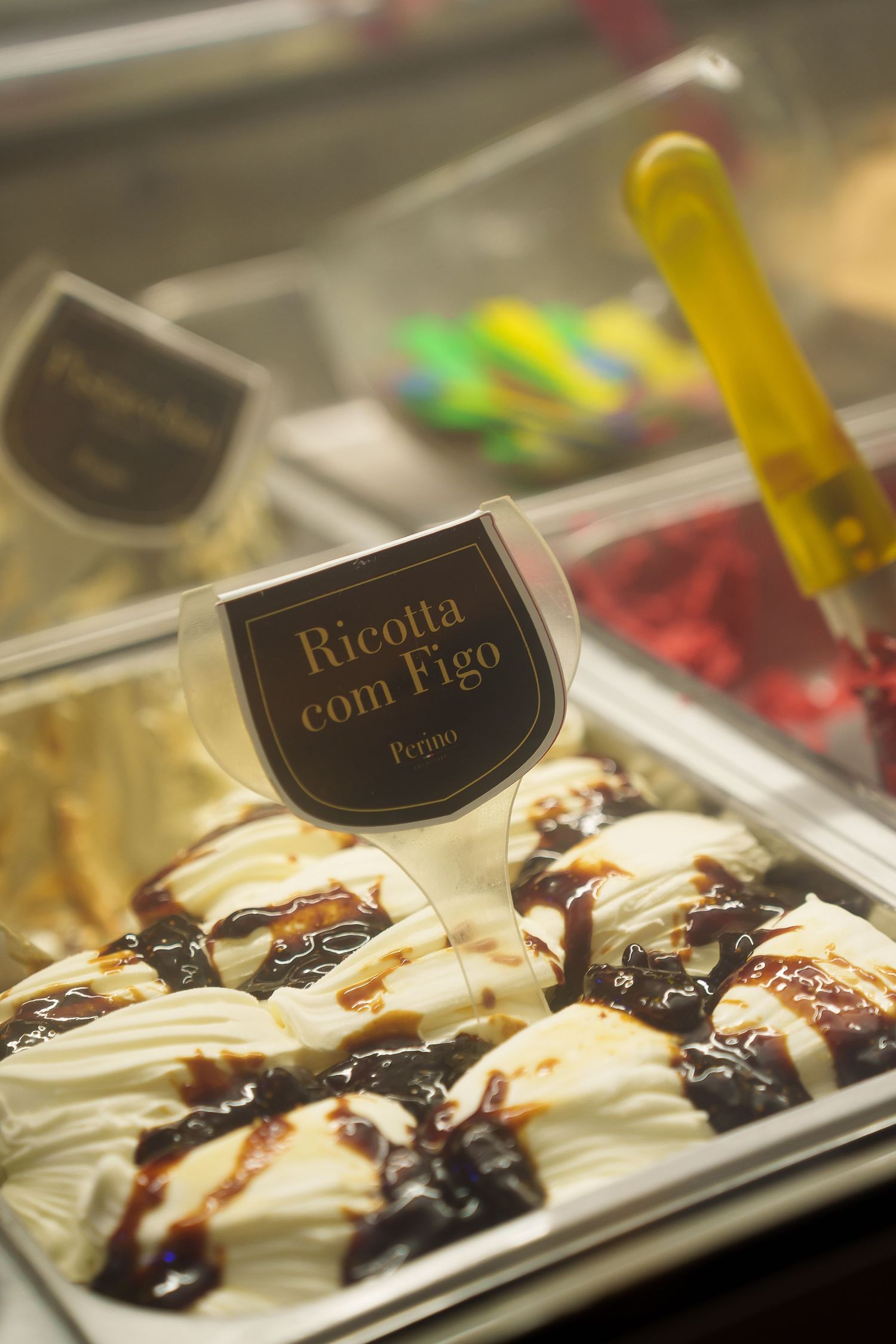 Ricotta com Figo gelato displayed in a glass case, topped with chocolate sauce and fig jam at an Italian gelateria.