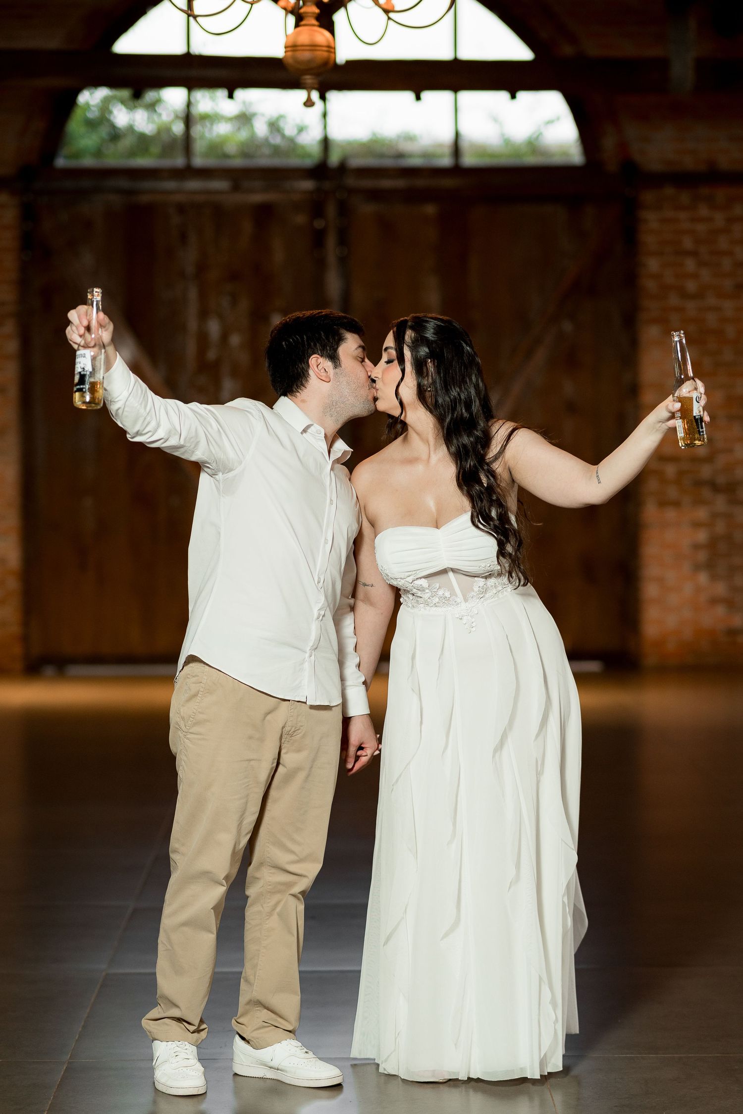 A couple in wedding attire kisses while raising beer bottles in a rustic barn venue.