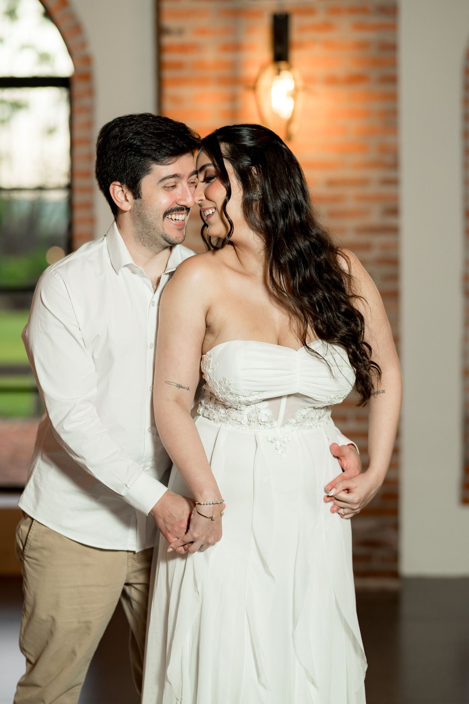 Couple in white outfits sharing a romantic moment, smiling and holding hands in a warmly lit brick venue.