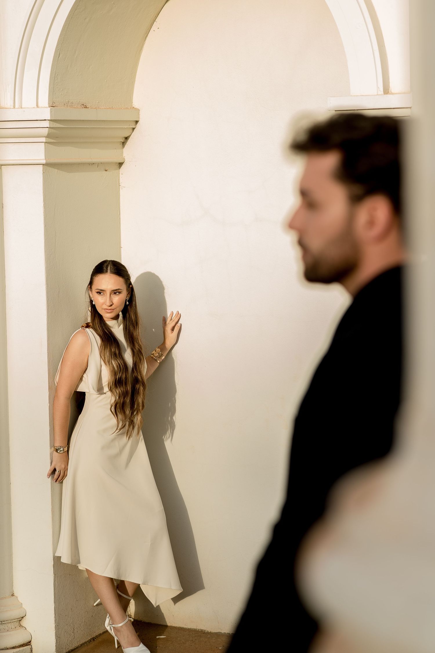 Elegant woman in beige midi dress leaning against white arch column while man in black suit stands in foreground.