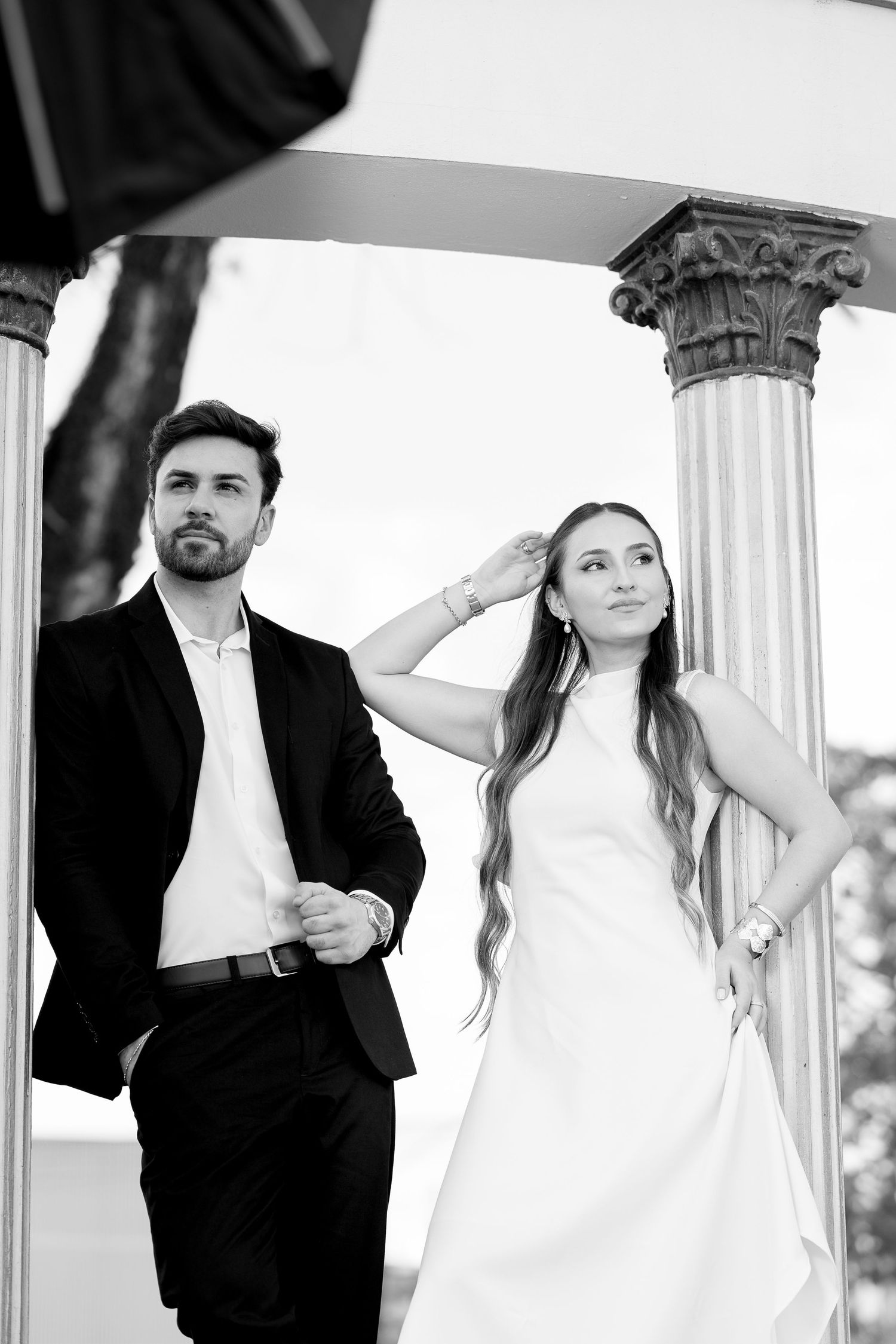 Black and white photo of elegant bride and groom posing between classical columns outdoors.