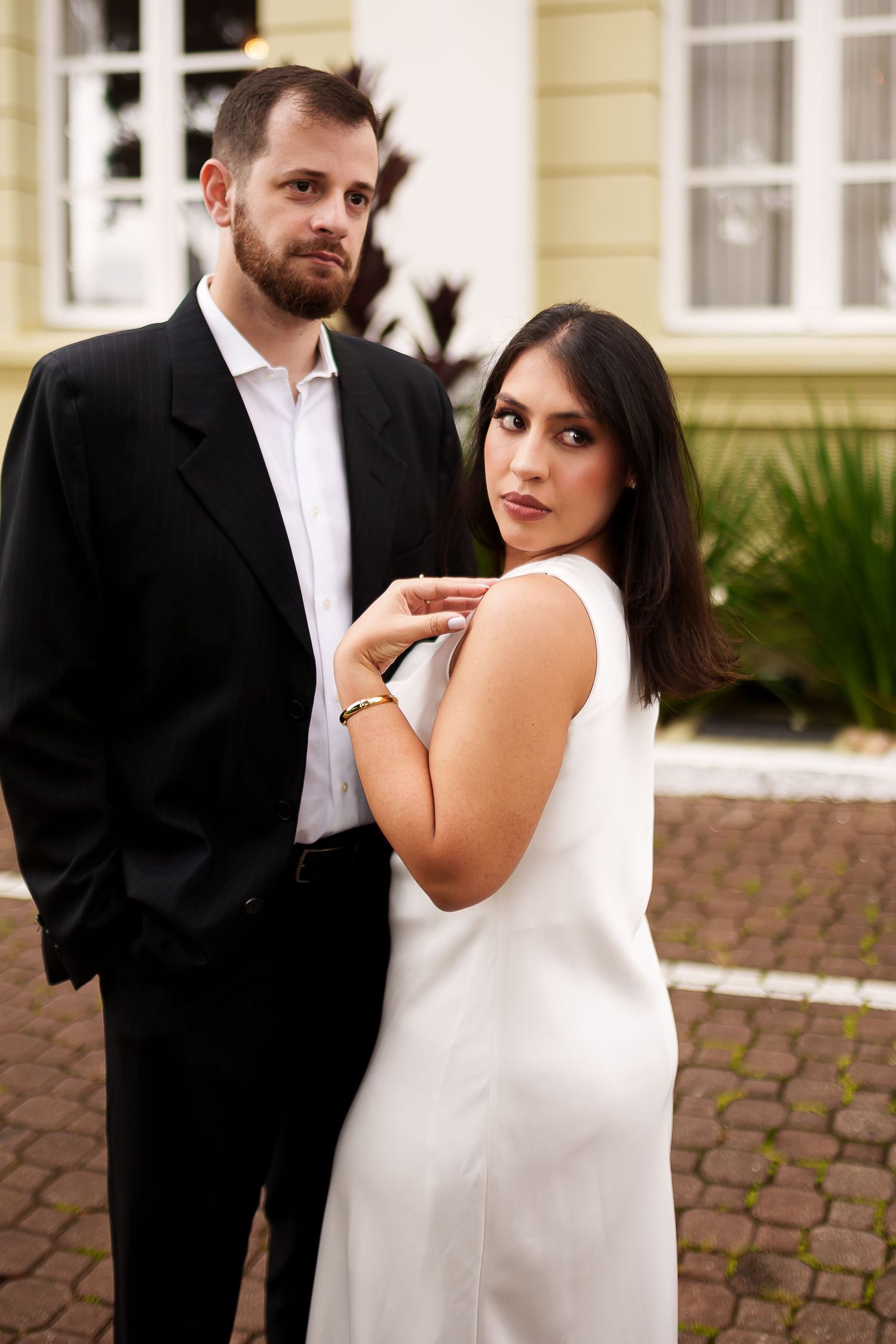 Elegant couple posing outdoors, man in black suit and woman in white dress, standing near a building.