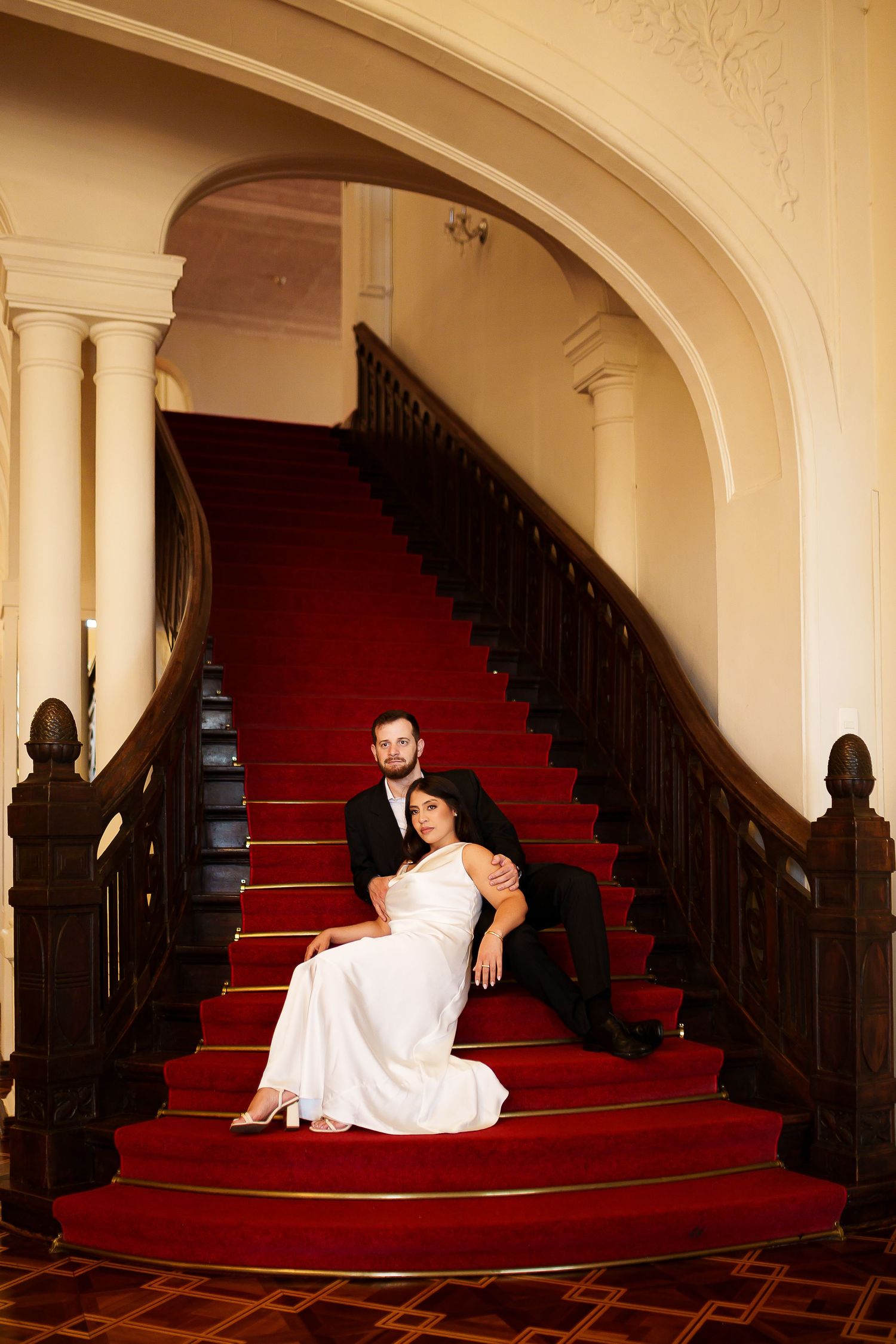 Bride and groom posing on a grand red-carpeted staircase with dark wood railings in an elegant venue.