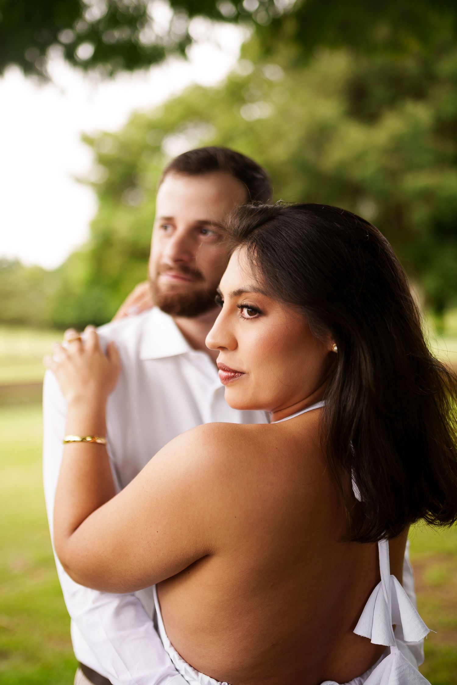 A couple embraces outdoors in a park, woman in white dress looks back over shoulder, man with beard behind her.