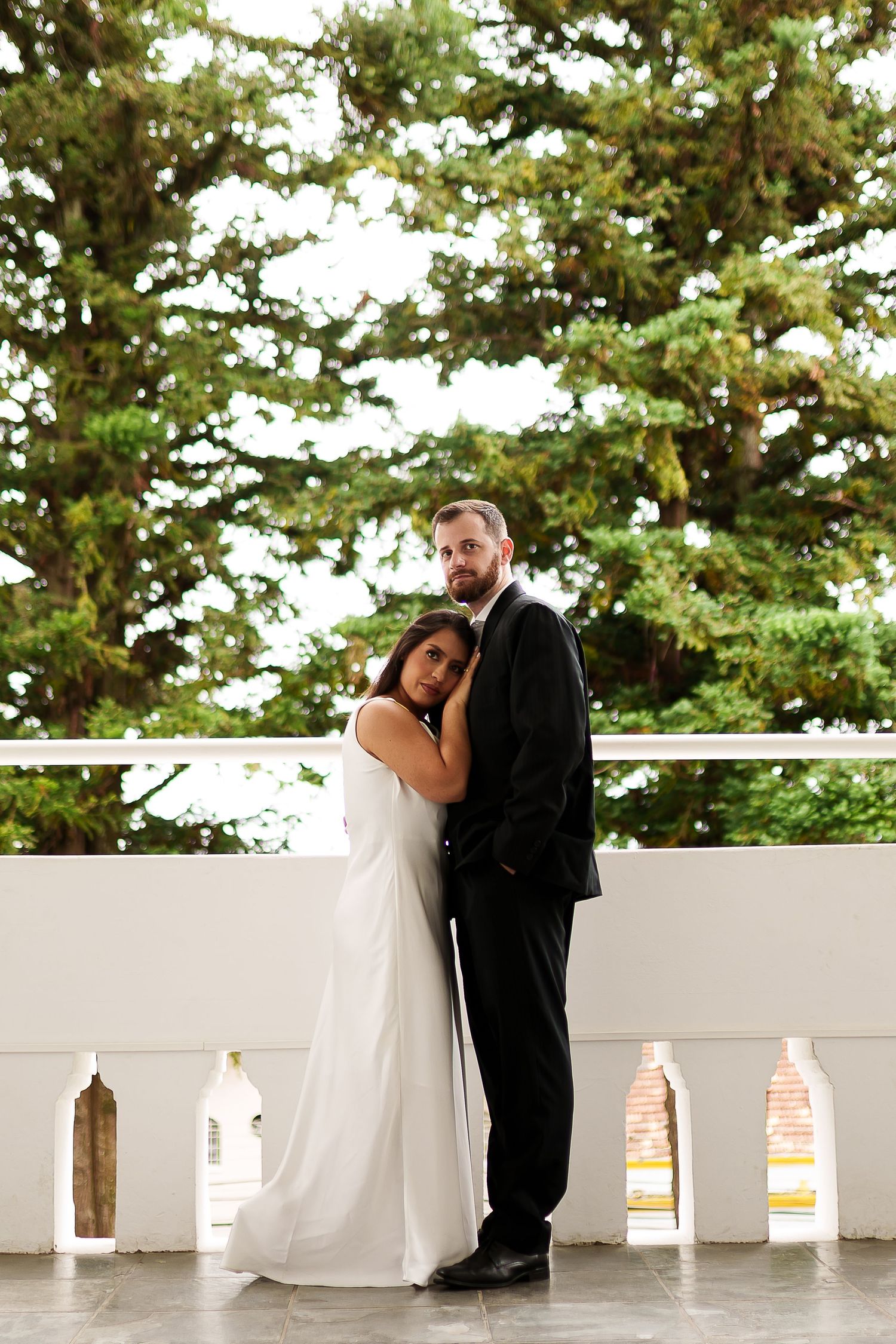 Bride in white gown leans against groom in black suit on a white balcony with lush green trees behind them.
