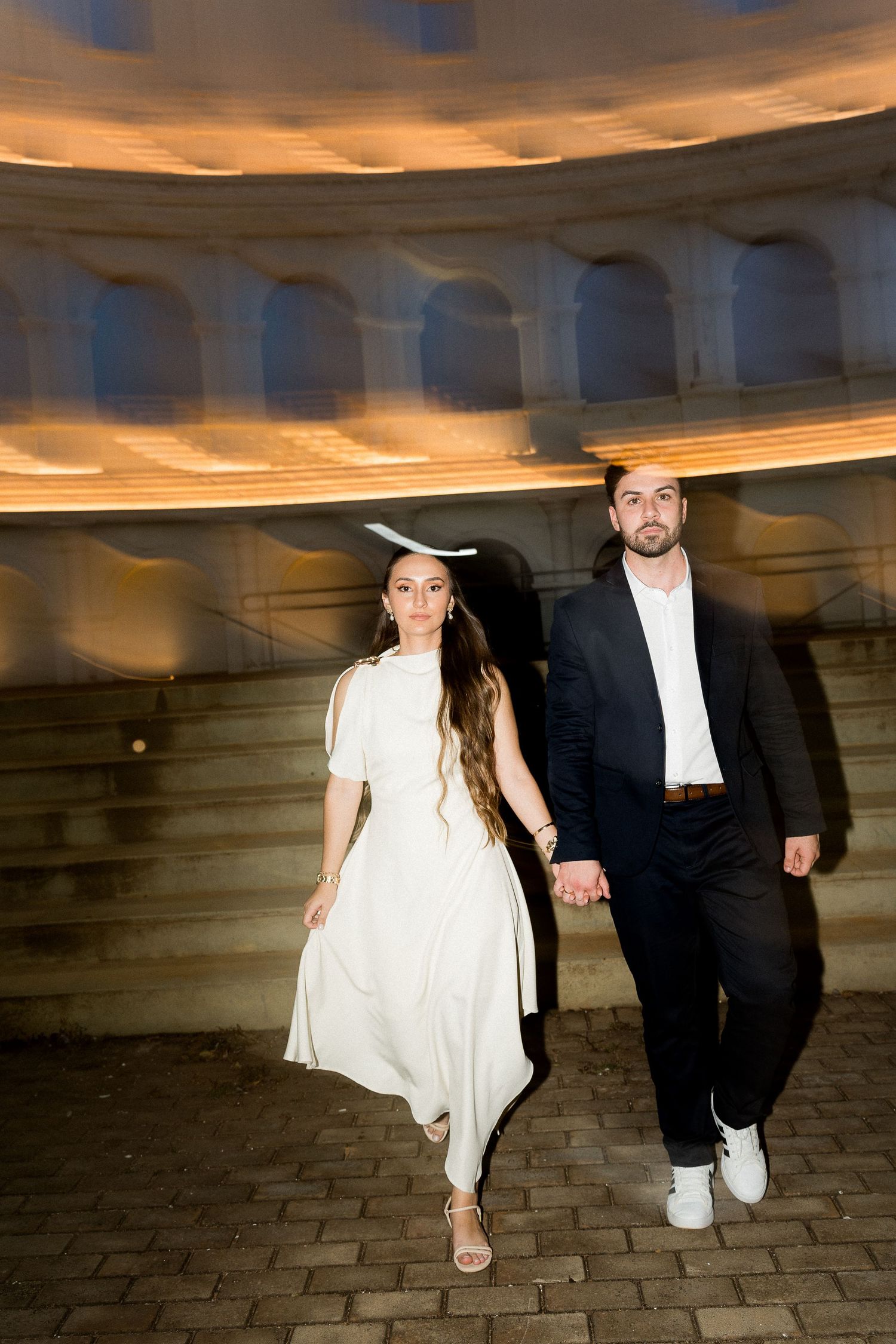 Couple in formal attire holding hands walking under an illuminated archway at night.