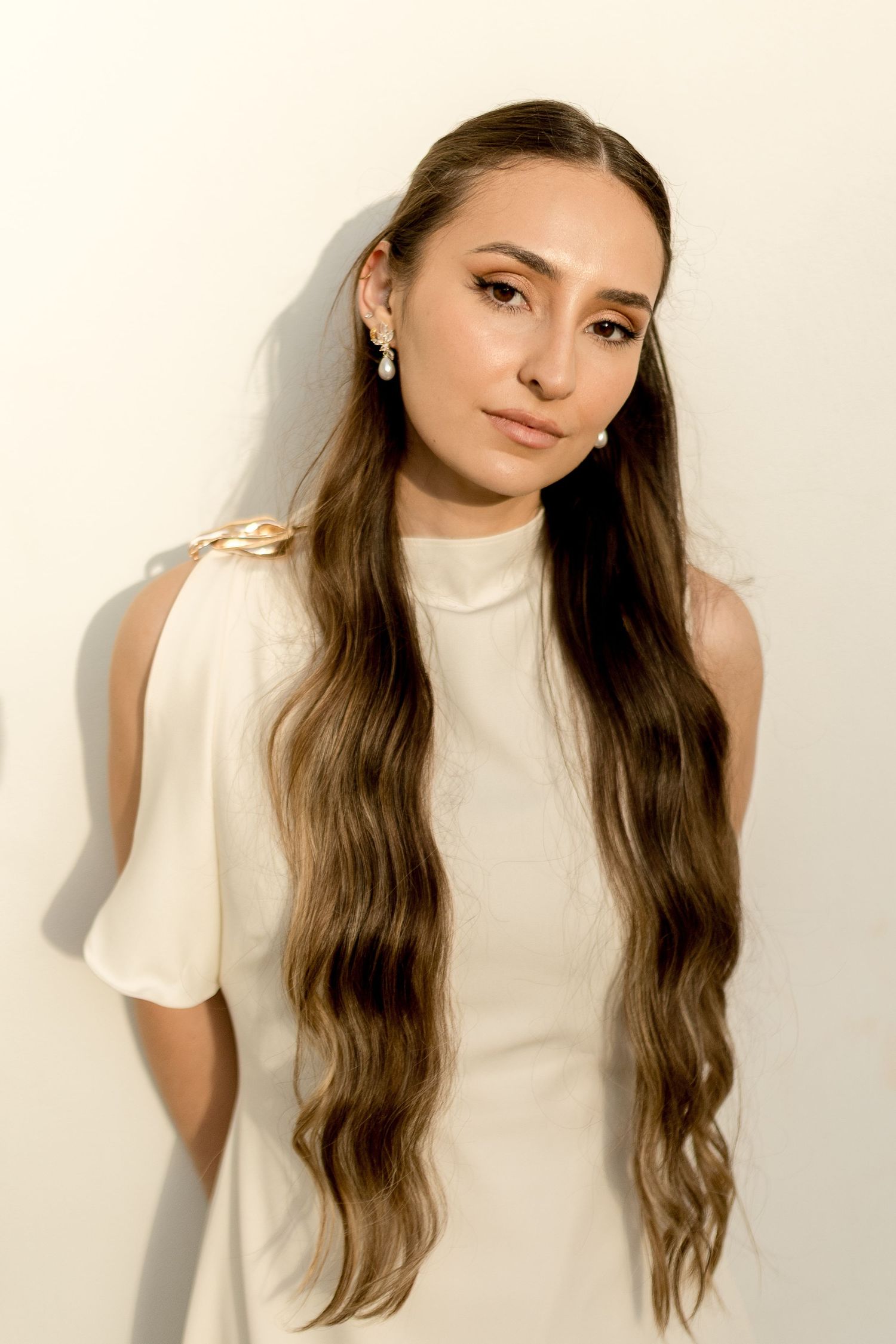Young woman with long wavy brown hair wearing a white dress and pearl earrings poses against a white wall.