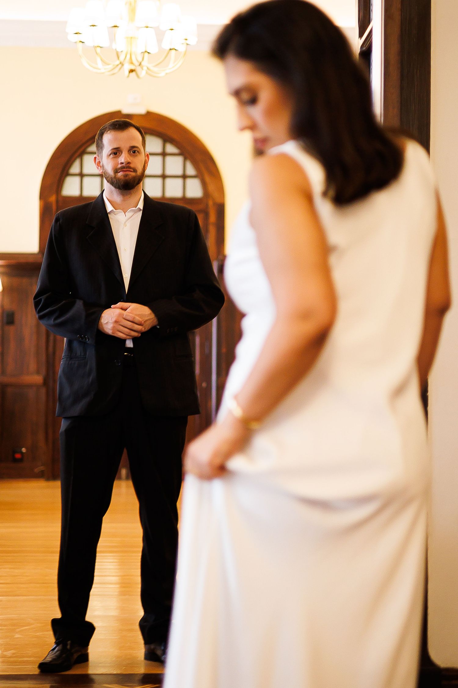 Groom in black suit watches bride in white dress during wedding first look moment in elegant venue.