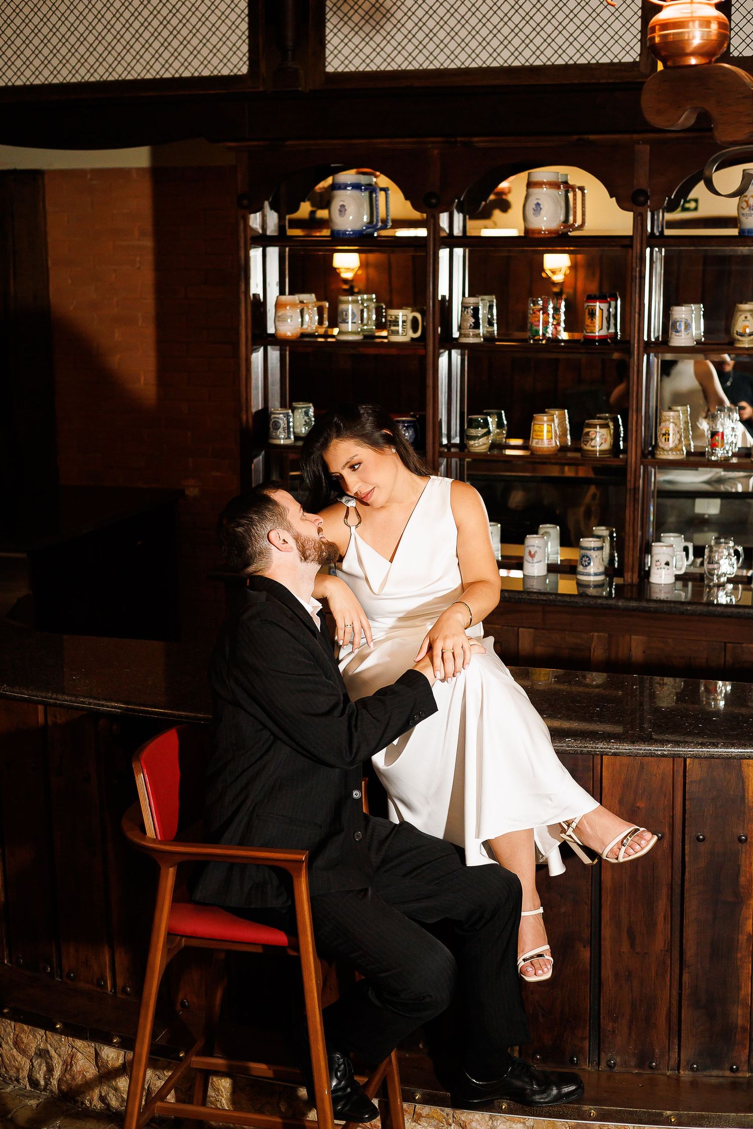 Couple sharing a romantic moment in a vintage bar, woman in white dress sitting on mans lap in black suit.