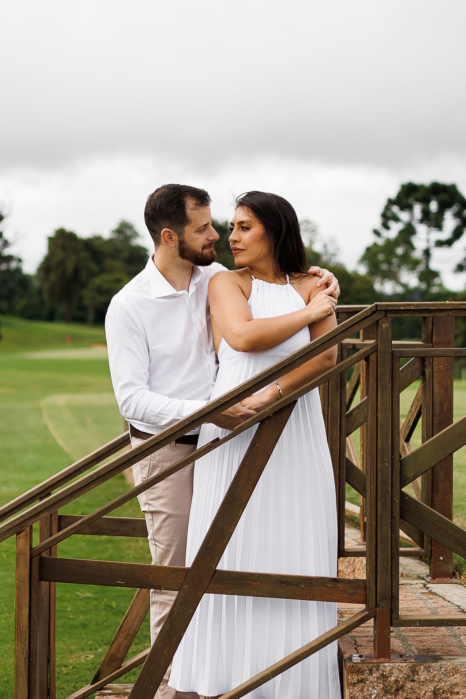 Romantic couple in white outfits embracing on a wooden bridge railing with lush green golf course background.