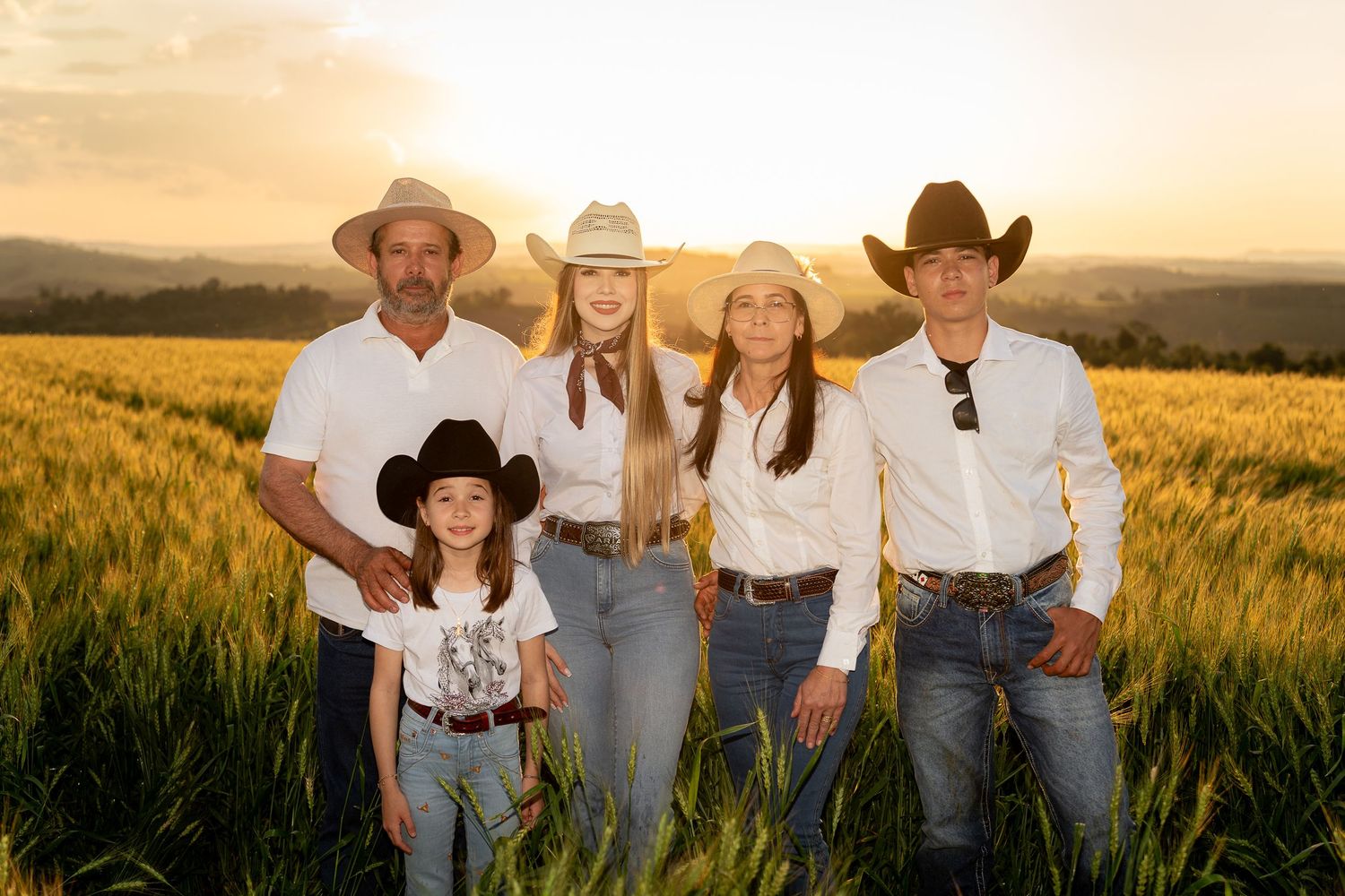 Family of five in cowboy hats posing in golden wheat field at sunset wearing white shirts and jeans.