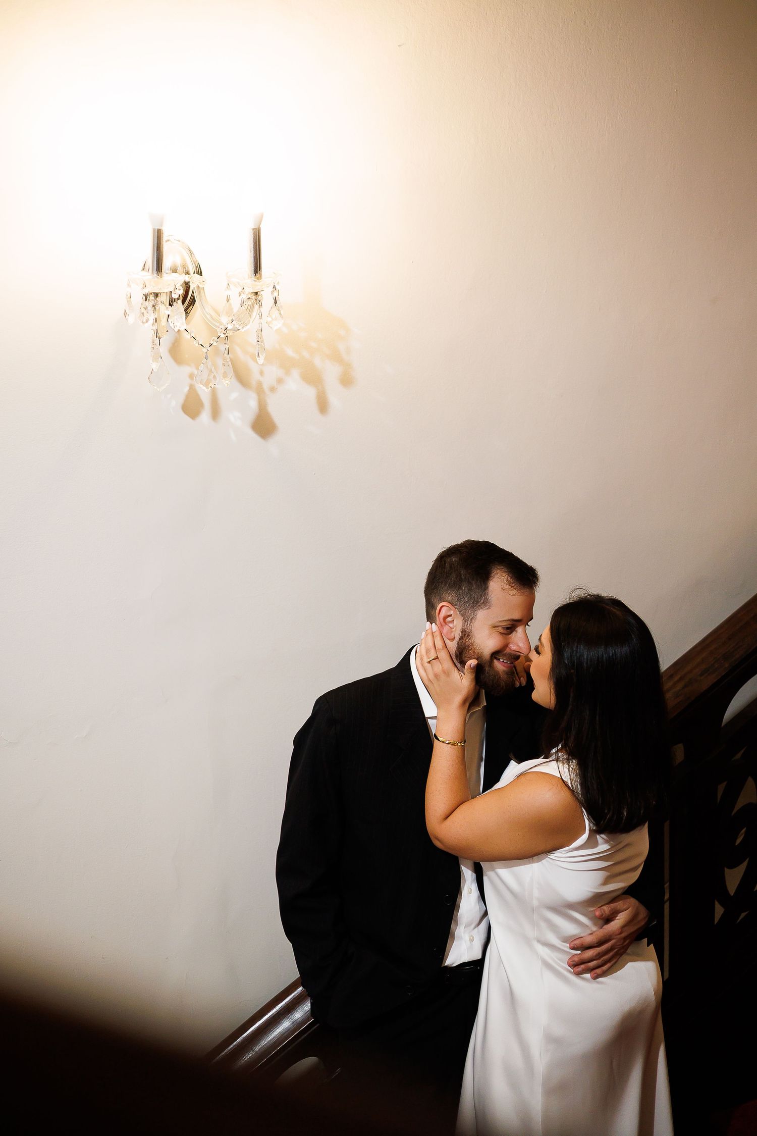 A couple shares a romantic kiss on a staircase beneath a crystal chandelier, dressed in wedding attire.