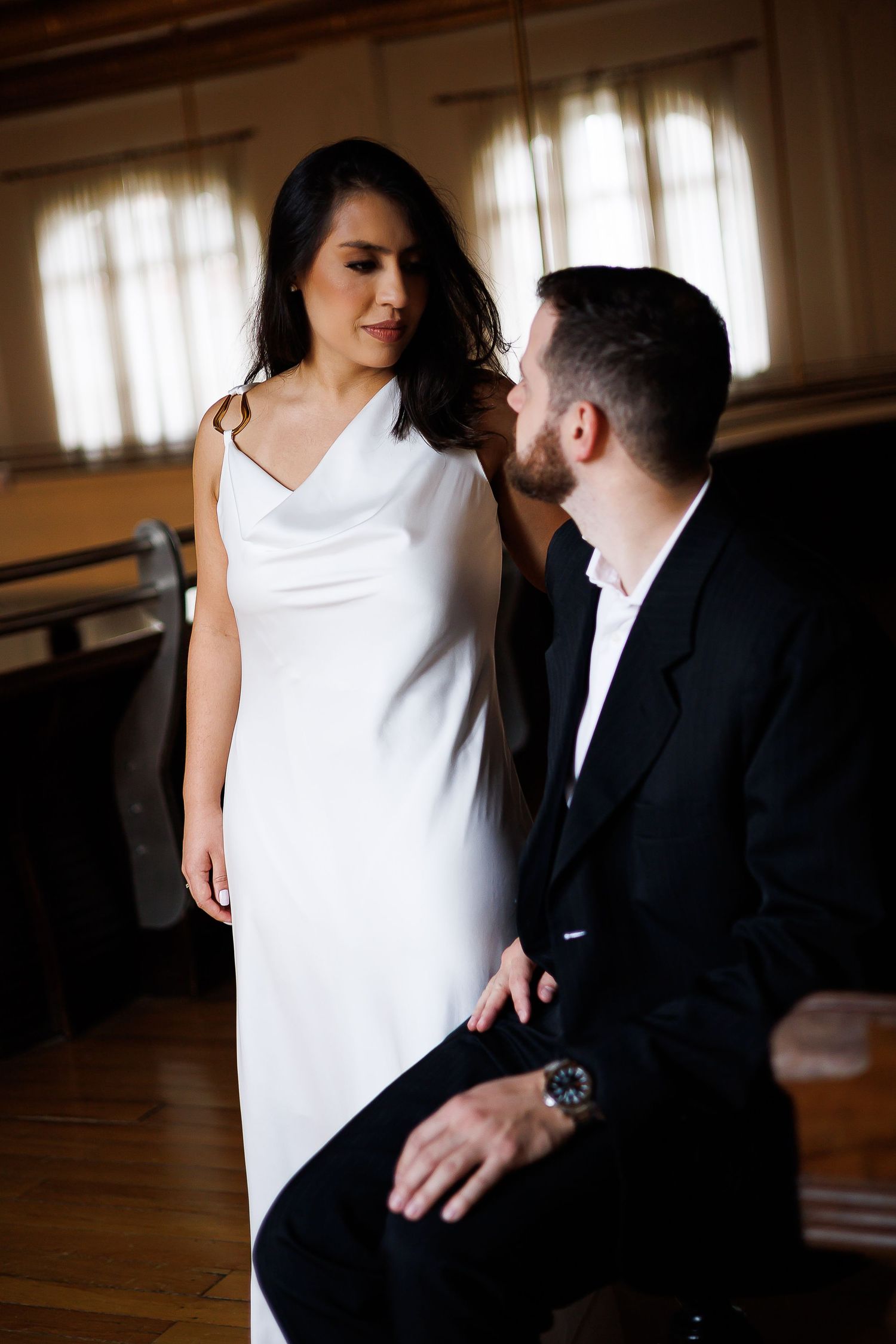 Bride in white cowl neck gown stands beside groom in black suit during intimate indoor engagement session.