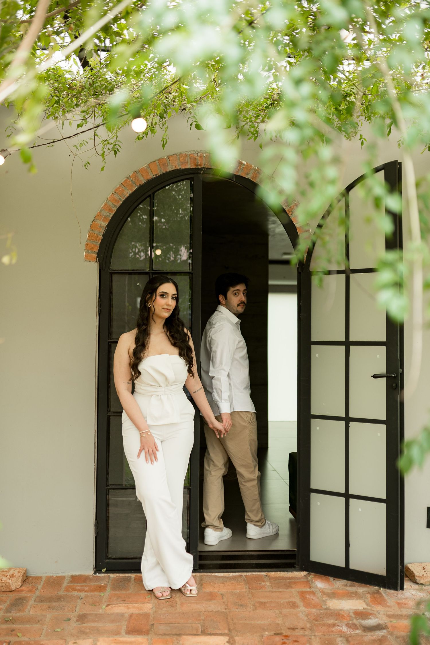 Couple in white and beige attire standing in an arched black French door surrounded by lush green foliage.
